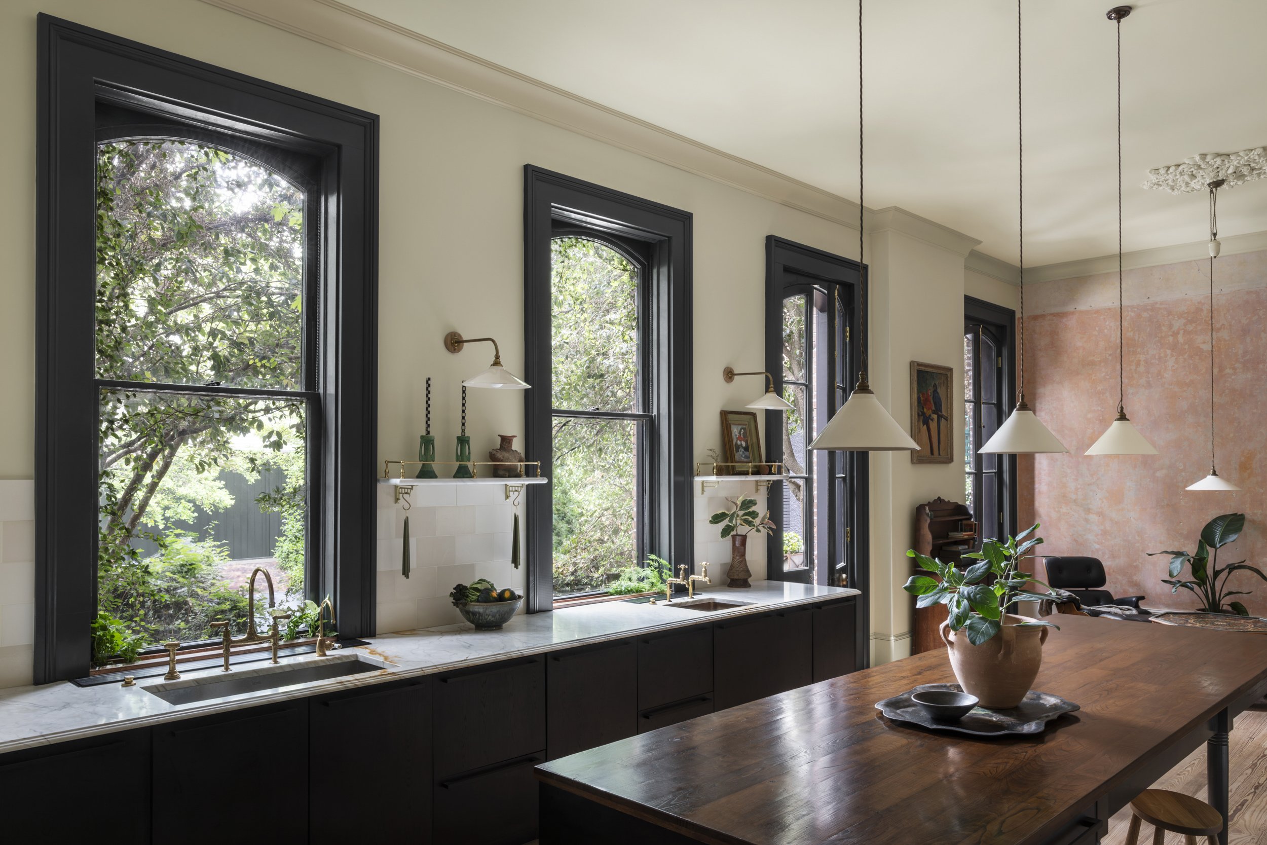 A bright kitchen with three large black-framed windows overlooking a garden with trees, decorated with plants, gold fixtures, and framed art. A marble countertop under the windows has a bowl of lemons, and a small shelf with vases and pottery. Pendan