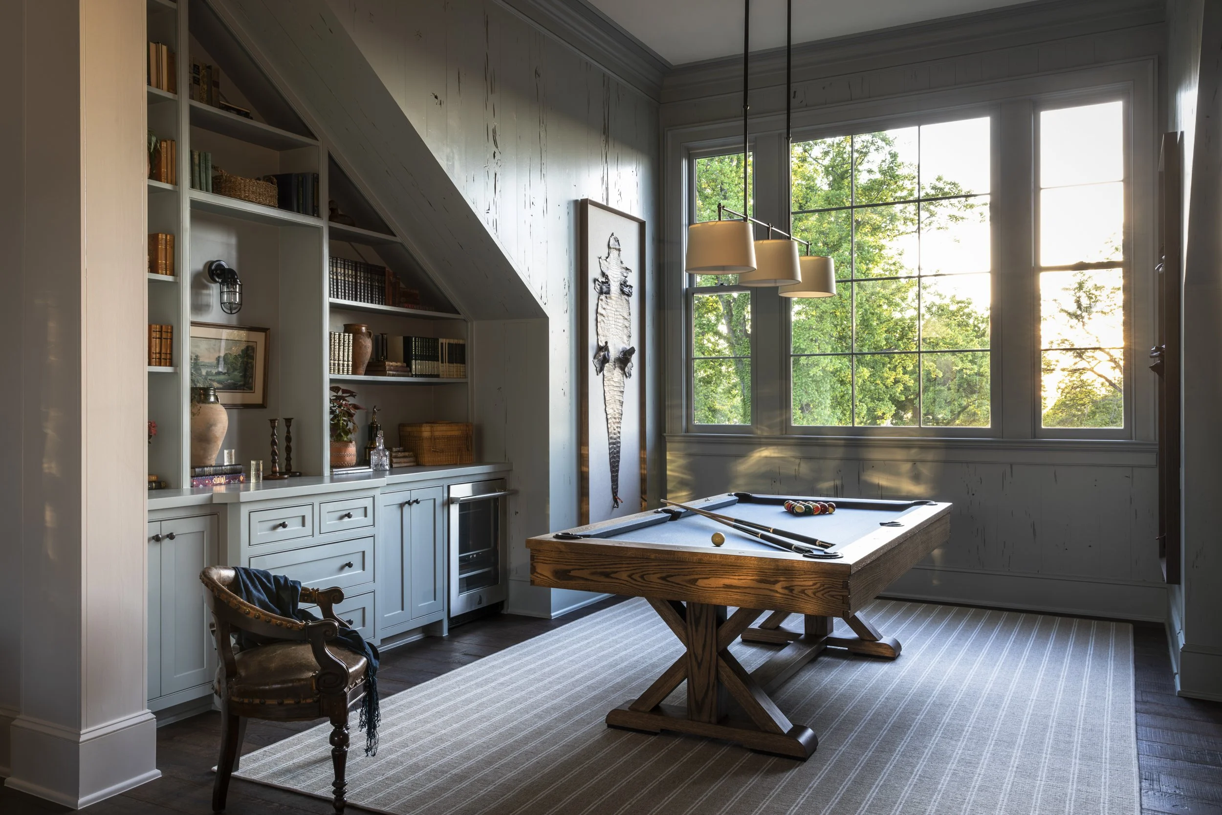 A room with large windows showing green trees outside, a billiards table with pool cues and balls, a built-in bookshelf with various items, and a hanging light fixture.