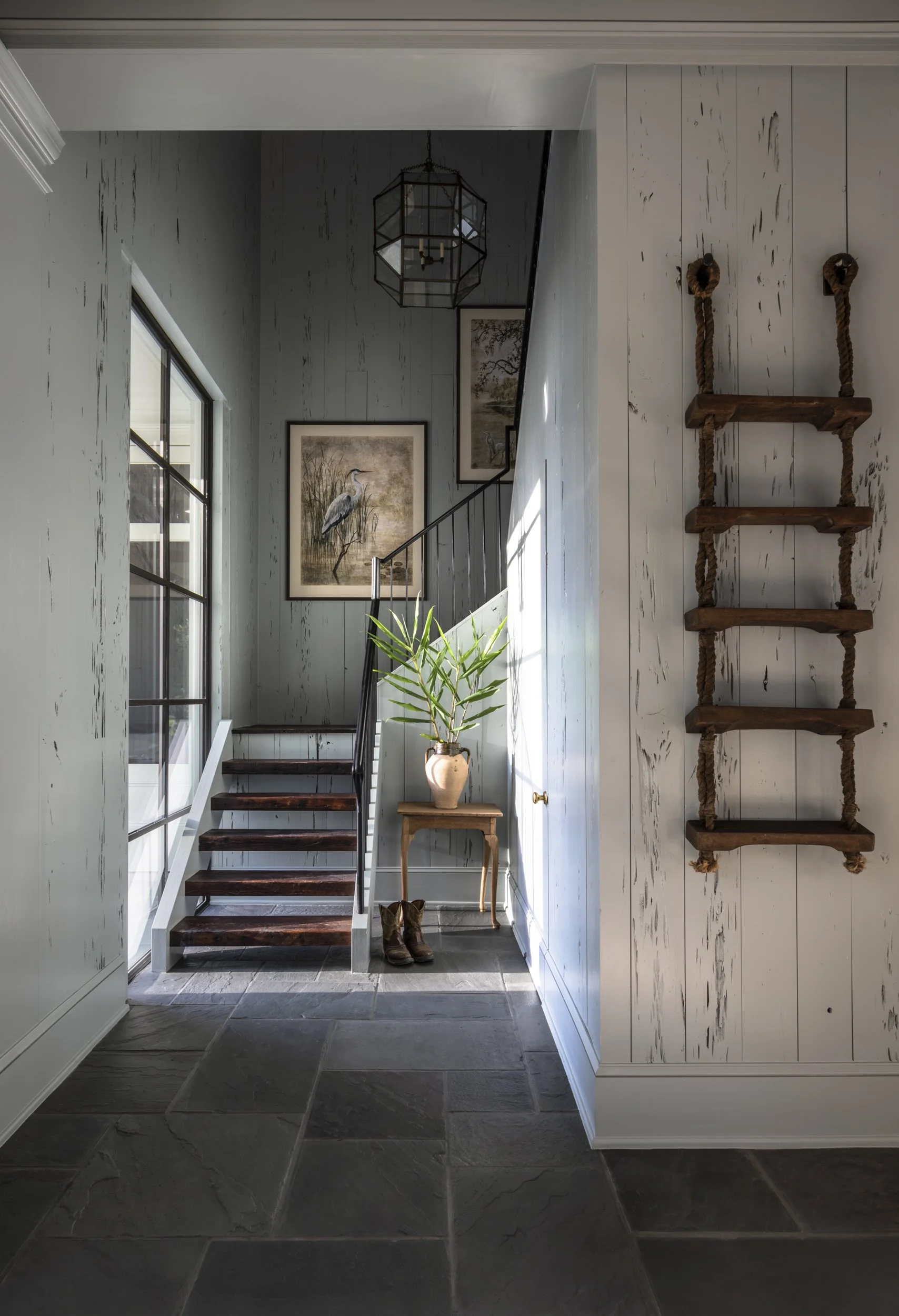 Interior view of a house entryway featuring dark wooden stairs with a black handrail, a potted plant on a small table, framed artwork on the wall, a rope ladder hanging on a white wooden panel, a large window with a grid of panes, gray slate tile flo