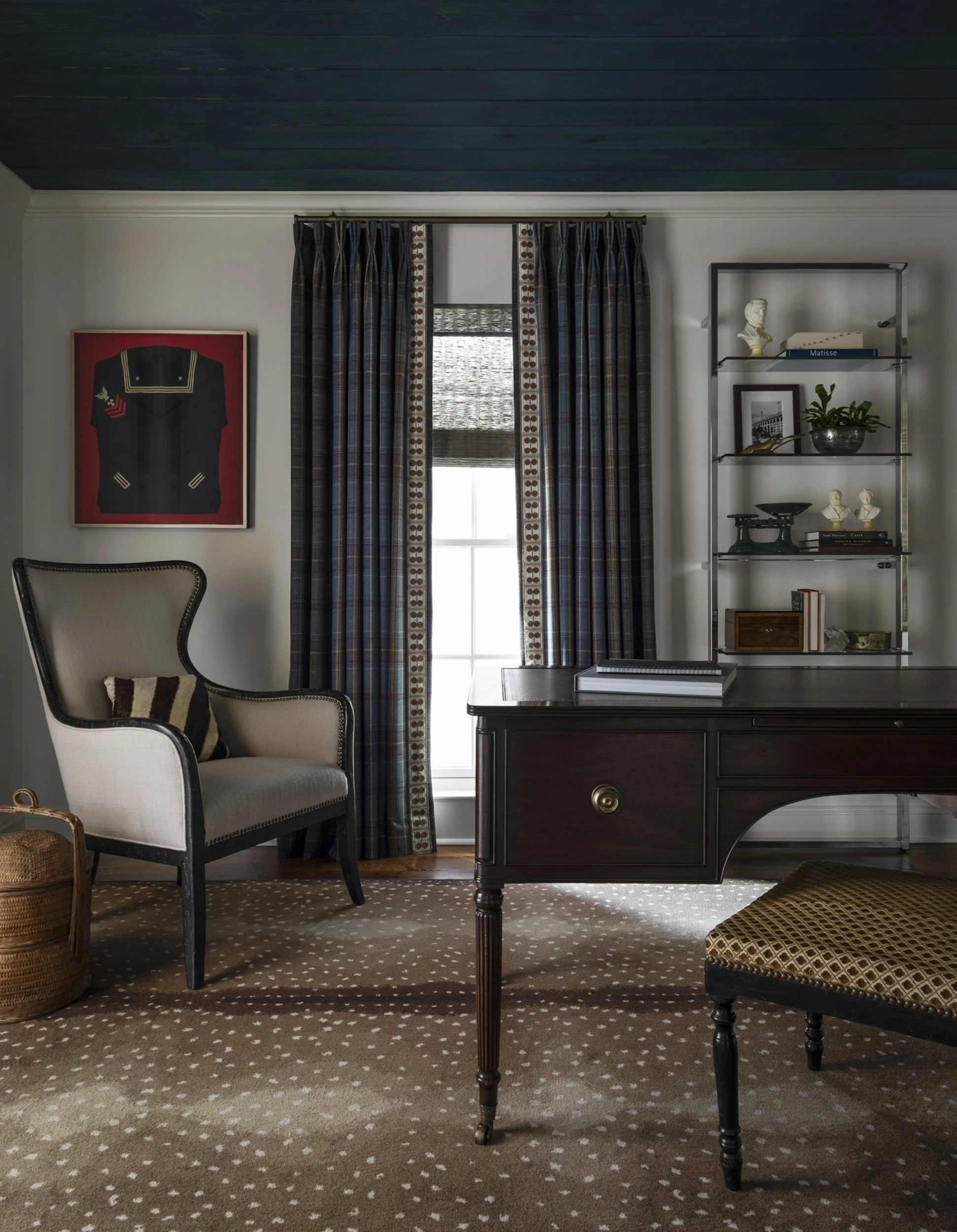 Interior of a room with a window with curtains, a framed portrait on the wall, a bookshelf with decorative items and books, a wooden desk with a closed book, and chairs with patterned cushions on a carpet with a polka dot pattern.