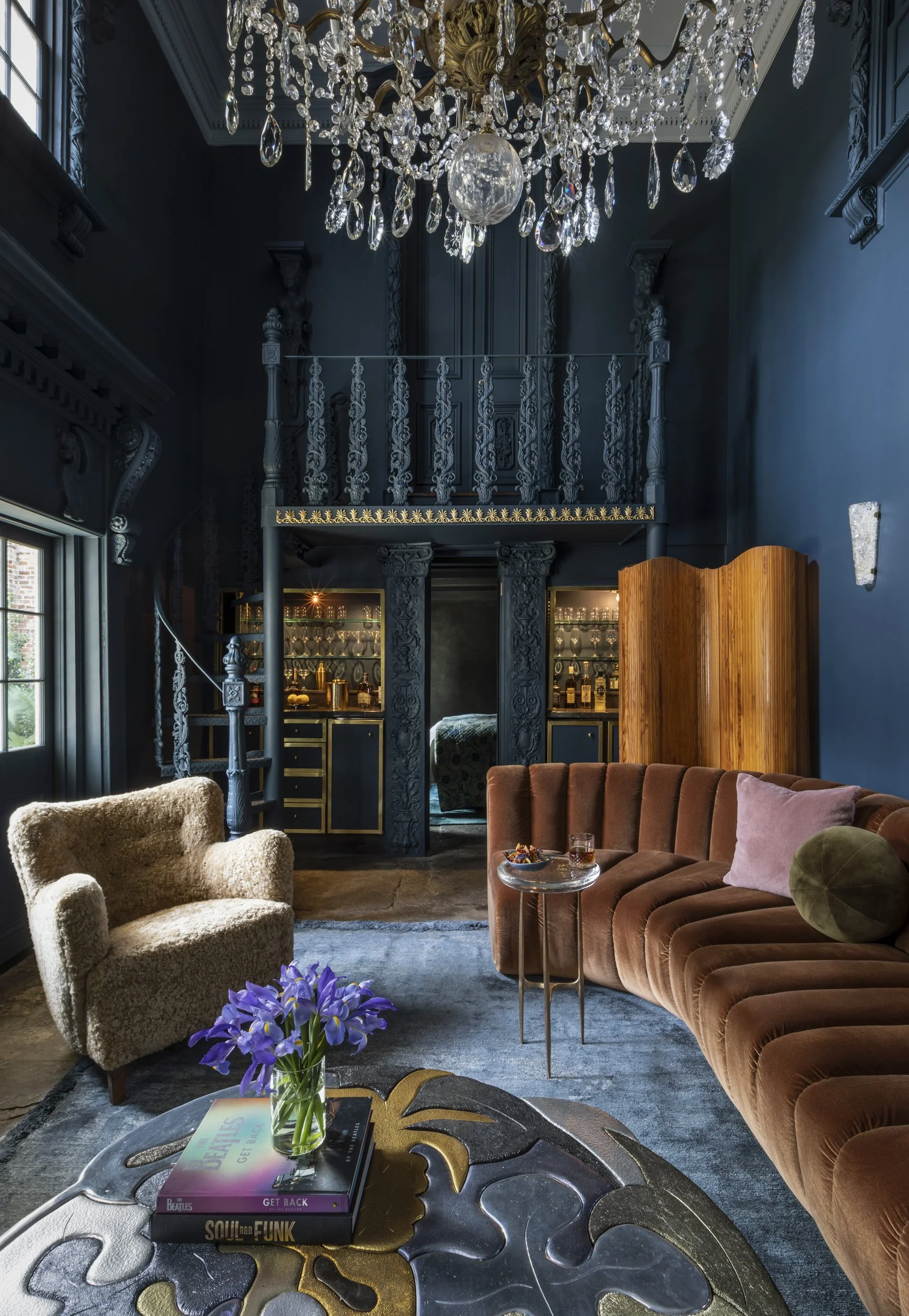 Elegant living room with a dark blue wall, ornate chandelier, curved velvet sofa, plush armchair, and a coffee table with magazines and a vase of purple flowers.