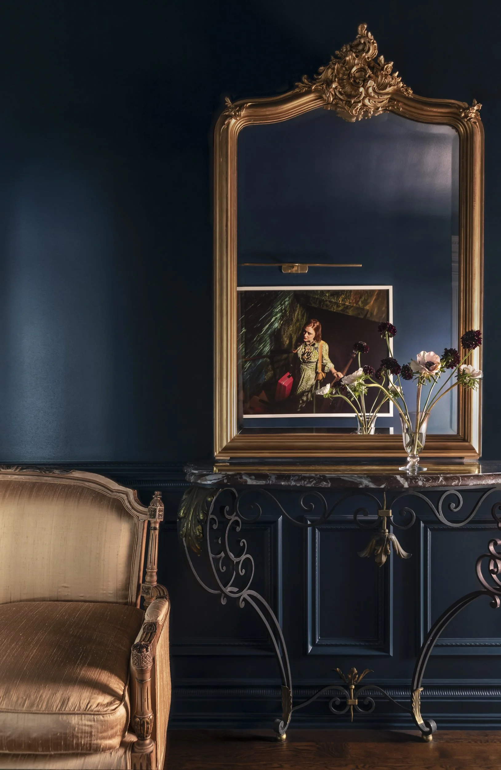 A decorative mirror with a gold ornate frame reflects a woman holding an umbrella and a red bag, standing in a room with dark blue walls and a black ornate table with a vase of flowers.