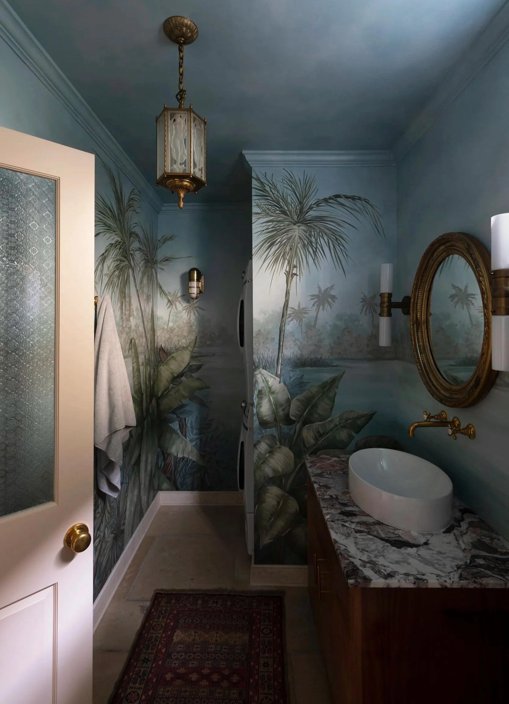 A bathroom with a painted mural of Palm trees on the walls, a white sink with a marble countertop, a gold framed mirror, a towel hanging, a ceiling with a hanging light fixture, and a small rug on the floor.