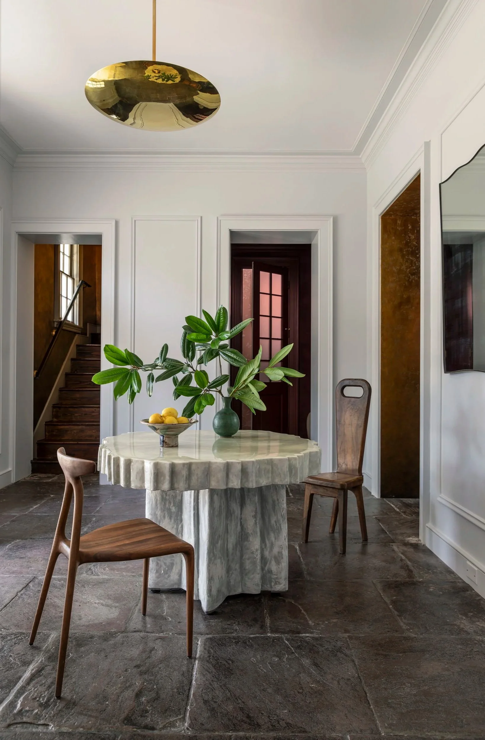 A dining area with a round table, two wooden chairs, a green vase with leafy plant, and a bowl of lemons. The room features white walls, decorative molding, dark stone flooring, a staircase with wooden steps, and an open doorway with a glass-paned do