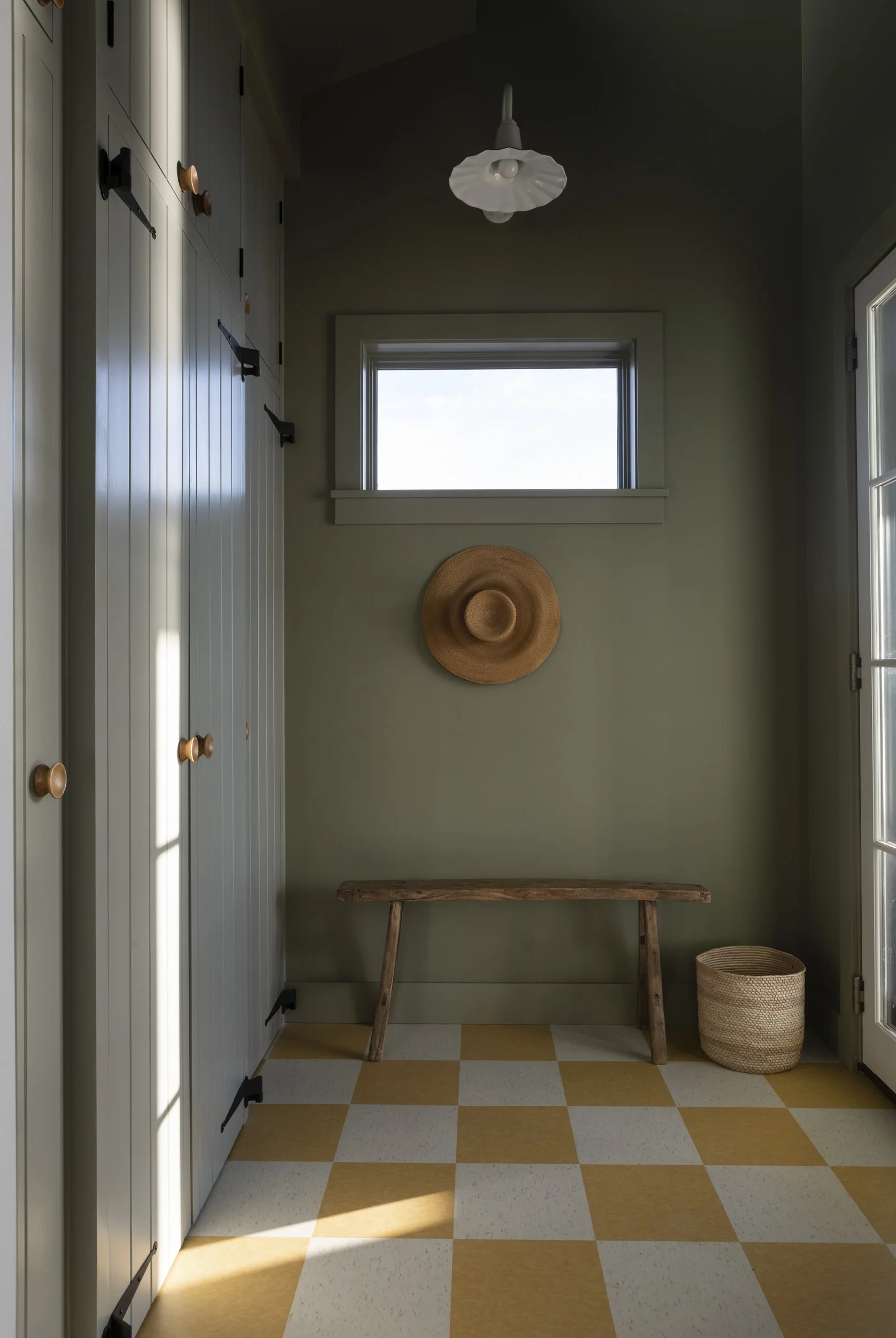 A small room with vertical paneled wall cabinets on the left, a window with natural light in the center of the back wall, a wooden bench beneath the window, a wide-brimmed straw hat hanging on the wall, a wicker basket on the floor to the right, and 