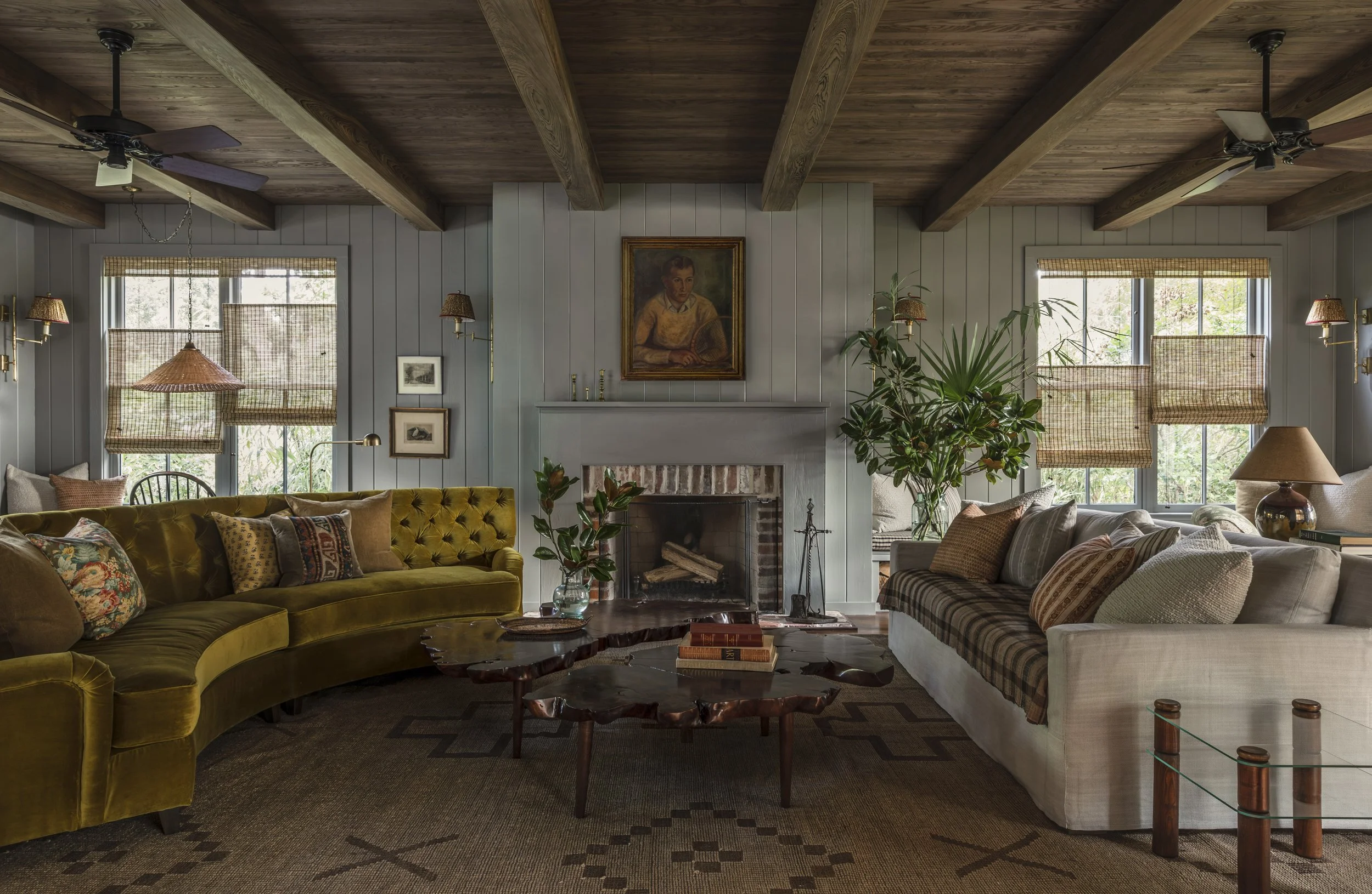 Living room with yellow velvet sofa, white sofa, wooden coffee table, and framed artwork above fireplace, with windows covered by woven shades, ceiling fans, and plants.