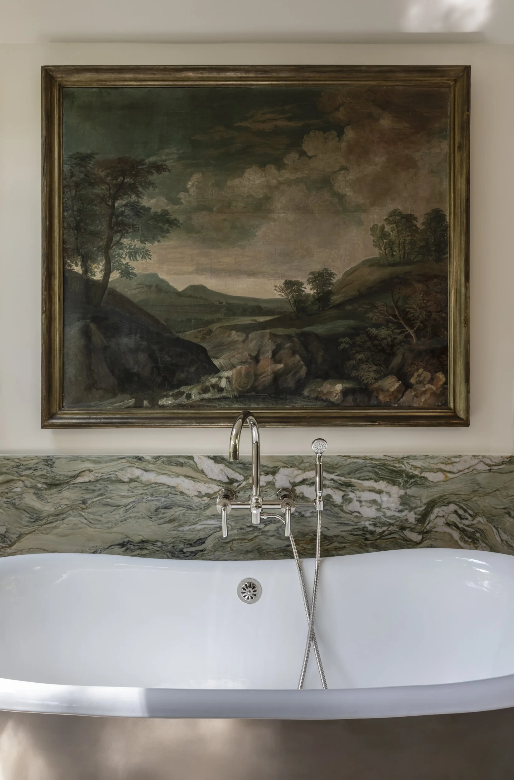 A bathroom sink with a silver faucet and shower attachment in front of a marble wall. Behind the sink is a framed landscape painting of a river, trees, and a cloudy sky.
