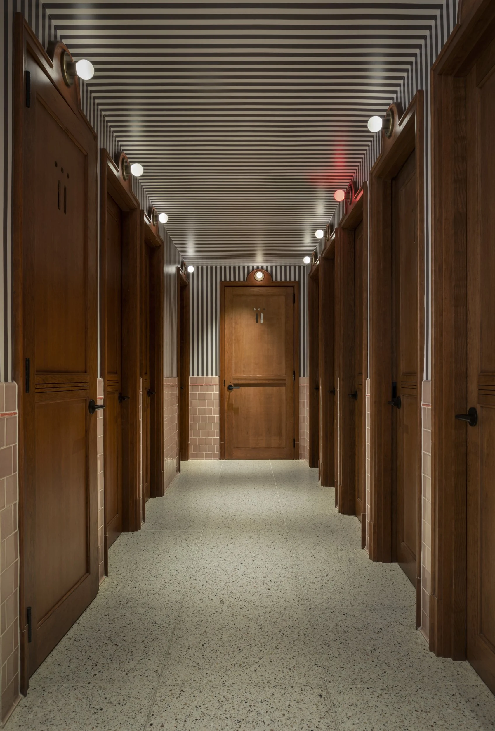 Communal bathroom corridor with wood stall doors, striped ceiling and globe occupancy lights at Virgin Hotels New Orleans