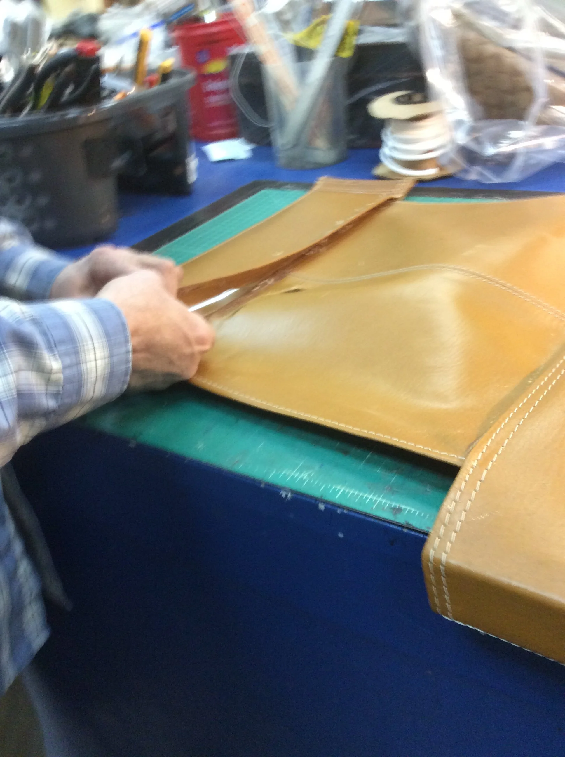 Close-up of a person working on a leather piece at a workbench, with tools and materials like spools of thread, a cutting mat, and a container of marking tools visible in the background.