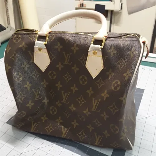 A Louis Vuitton handbag with the iconic monogram pattern, white handle, and gold hardware, placed on a tiled surface with office supplies in the background.