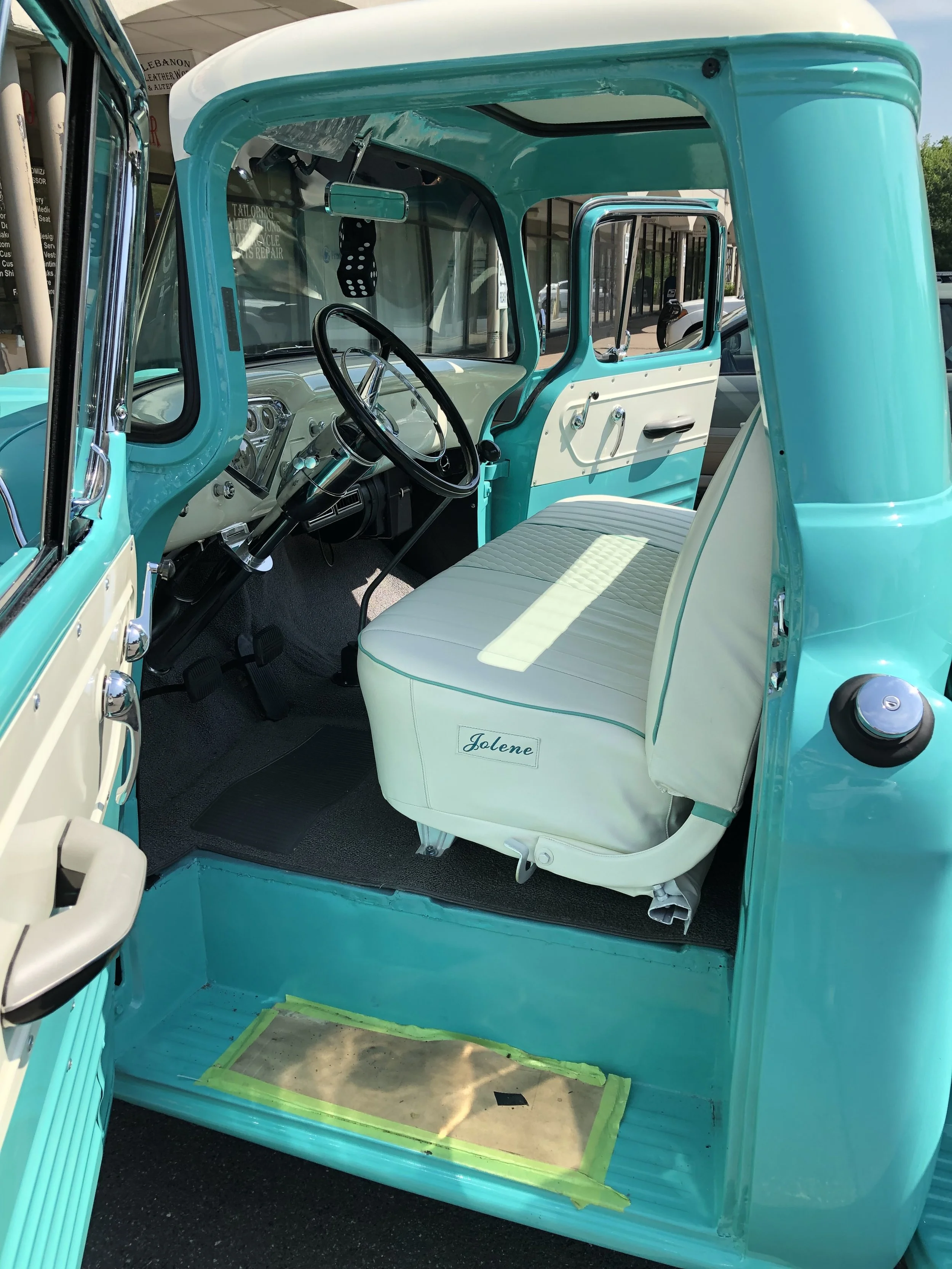 Interior of a vintage turquoise and white truck with a white bench seat, dashboard, and steering wheel, with the passenger side window open, showing a workspace or workshop environment outside.