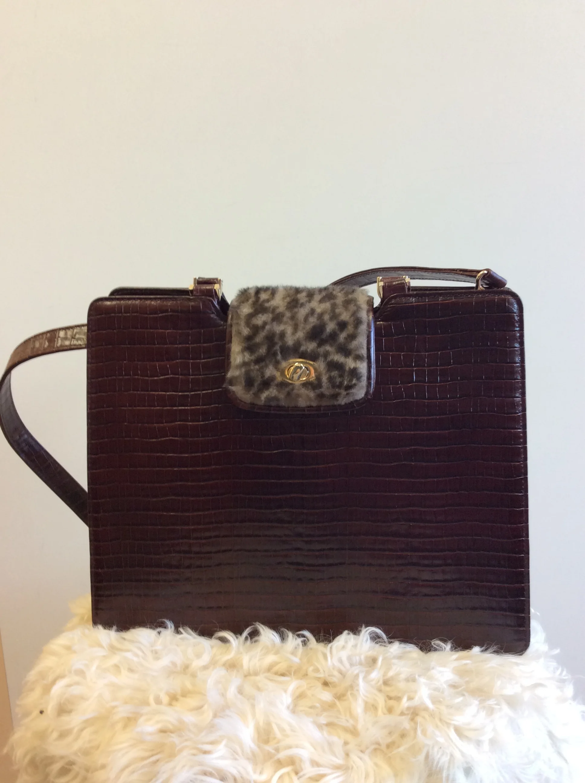 A brown crocodile-patterned handbag with a leopard print flap and gold clasp, placed on a fluffy white rug.