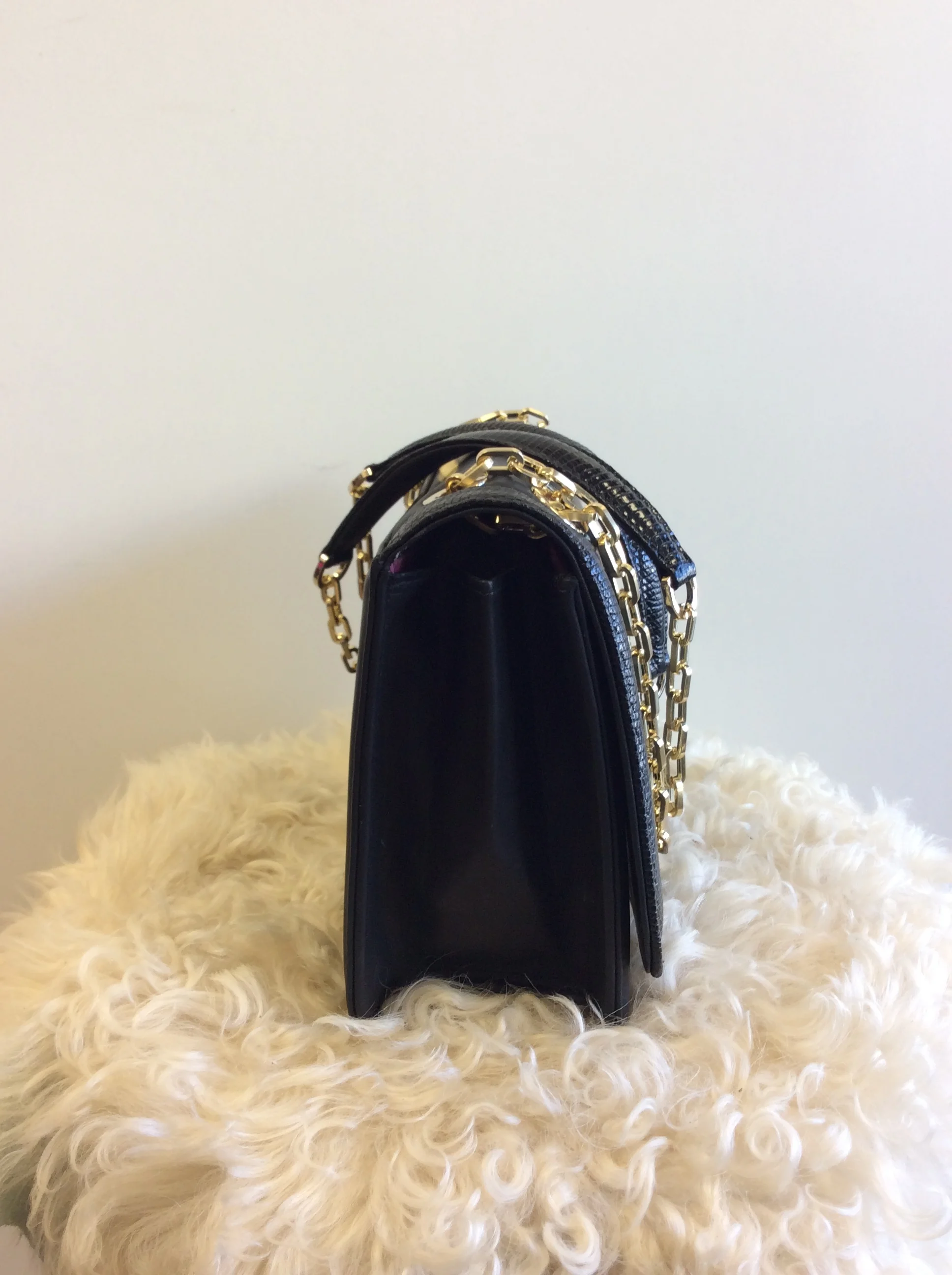 Black handbag with gold chain handles displayed on a fluffy cream-colored surface against a plain white background.
