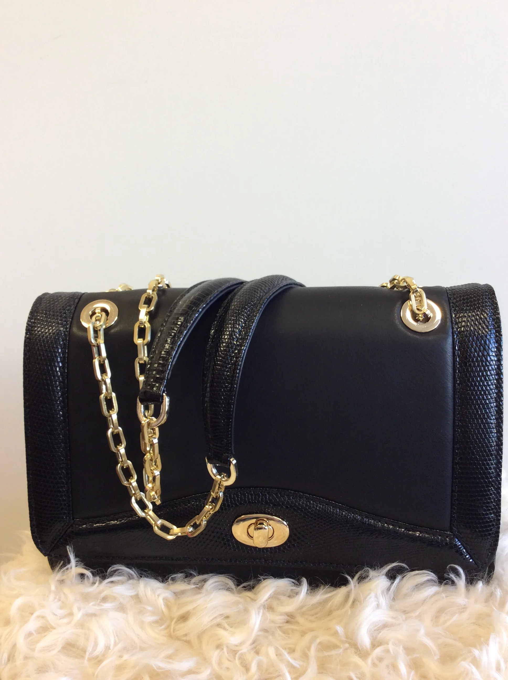 A black handbag with a gold chain strap and a gold turn-lock clasp, resting on a white furry surface.
