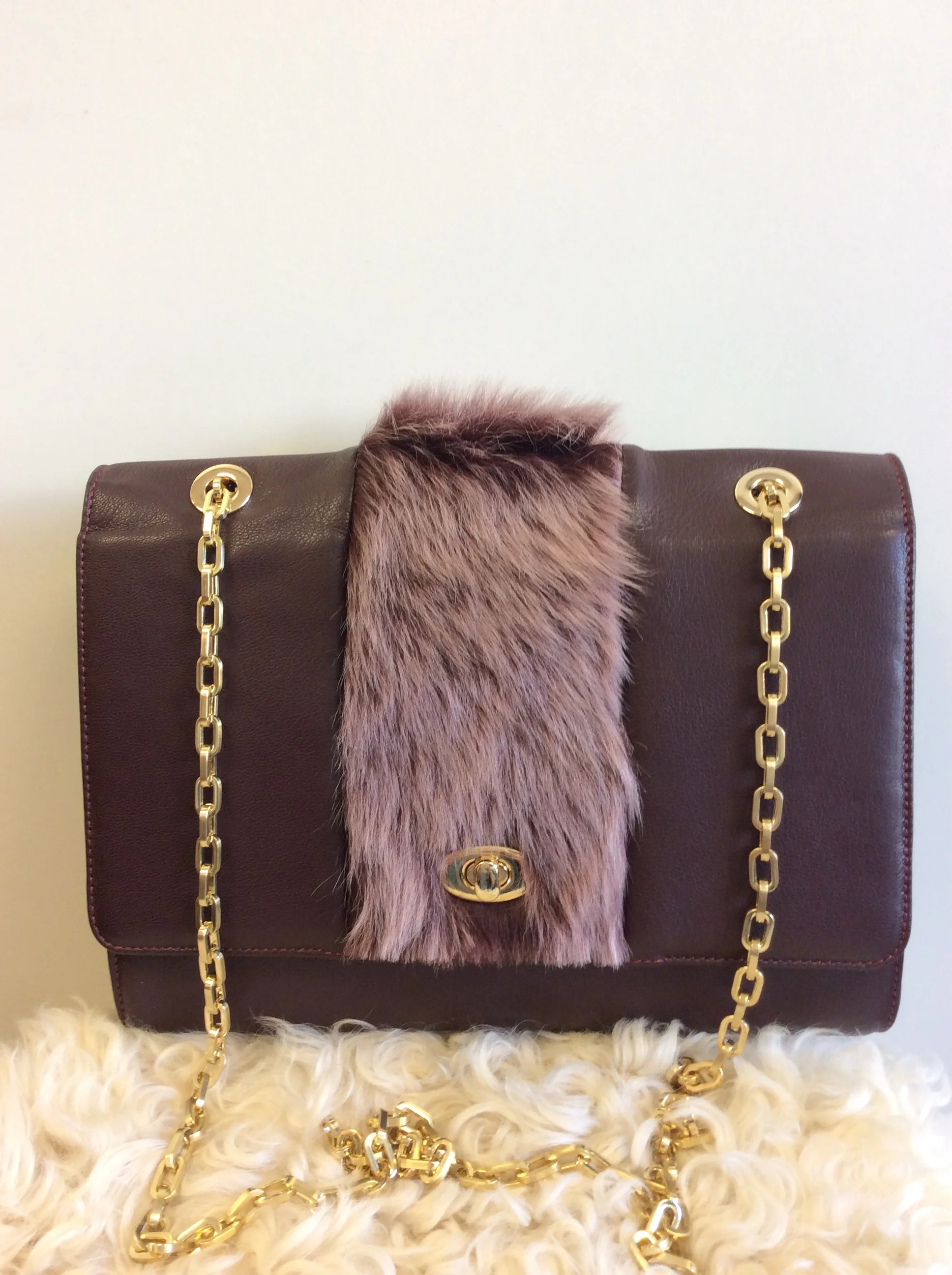 A brown handbag with a faux fur center strip, gold chain straps, and a gold clasp, placed on a fluffy white surface with gold chains around it.