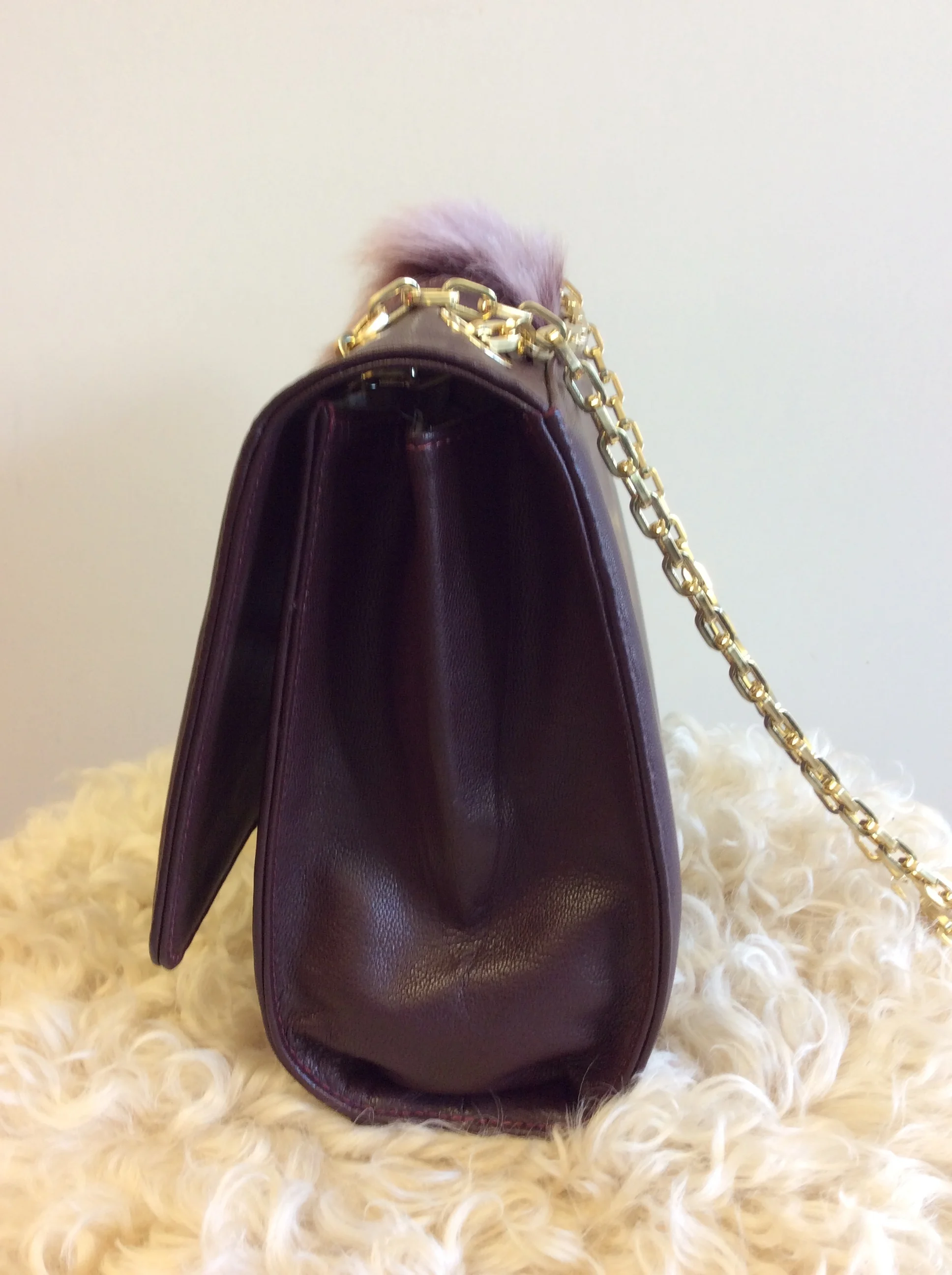 A burgundy leather handbag with a gold chain strap, resting on a soft, fluffy, cream-colored surface, with a plain, cream background.