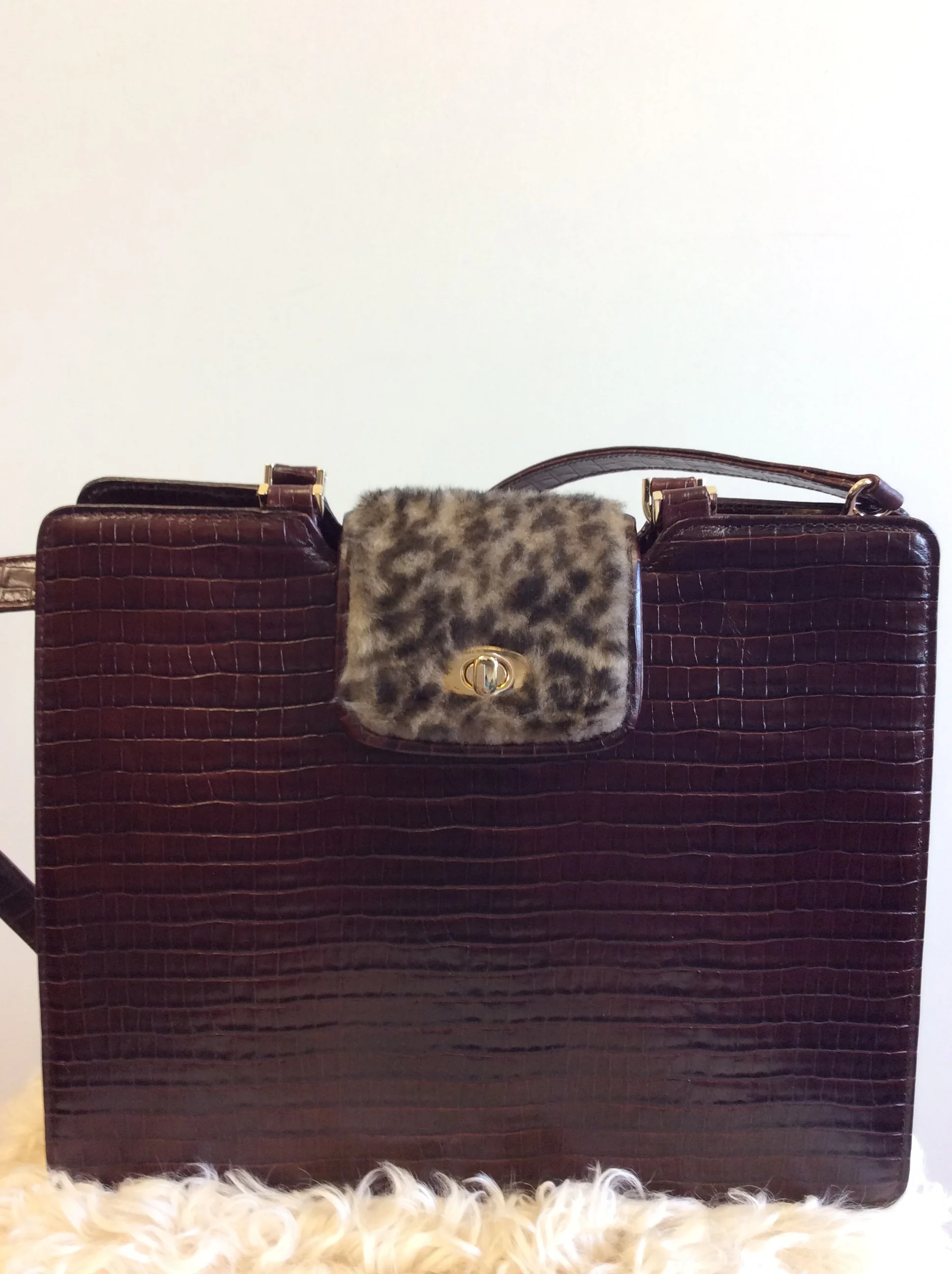 A dark brown crocodile-embossed leather handbag with a leopard print fur flap and gold hardware, placed on a light-colored furry rug.