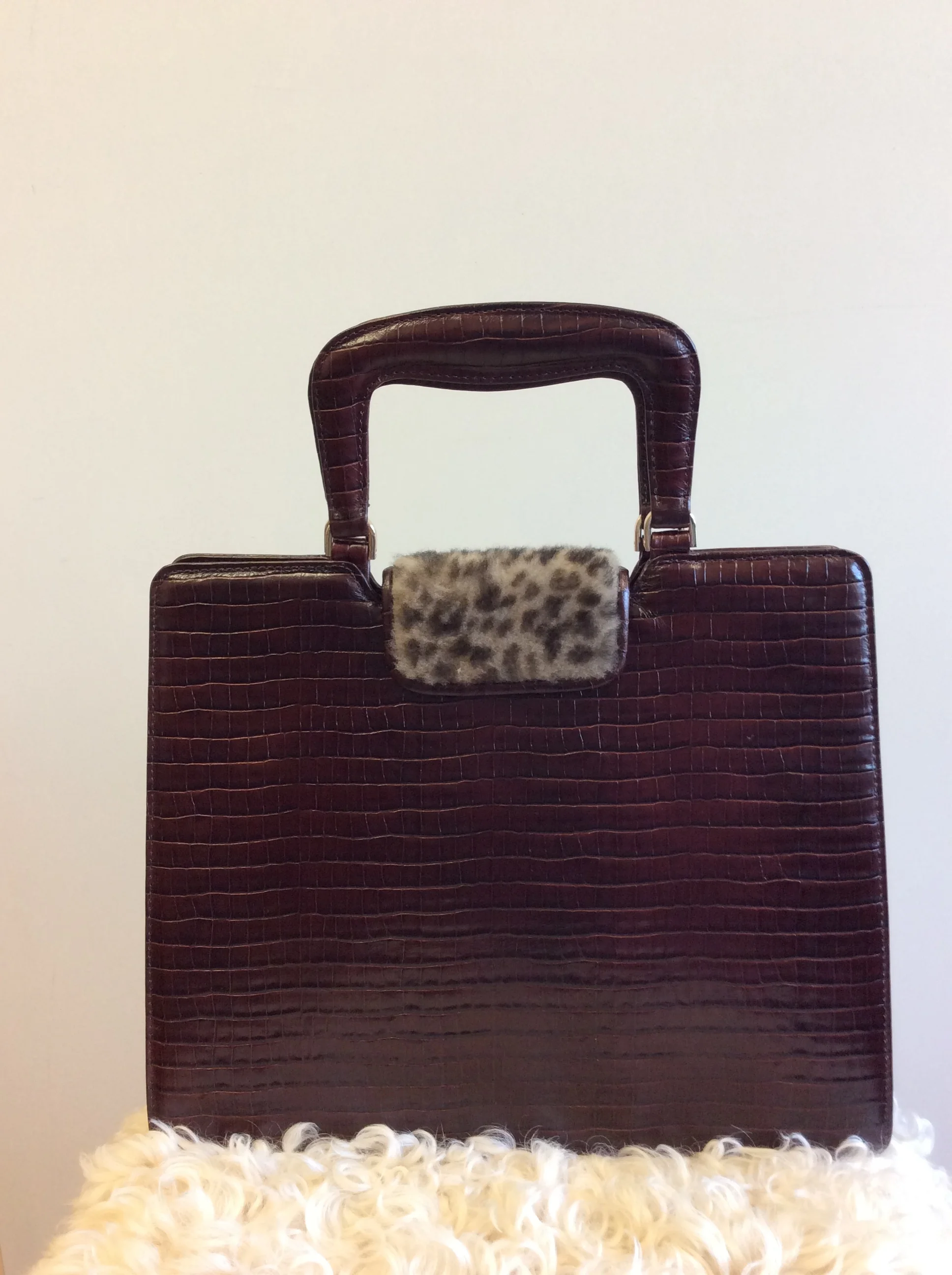 A vintage brown leather handbag with a leopard print scarf tied around the handle, placed on a white fluffy rug.