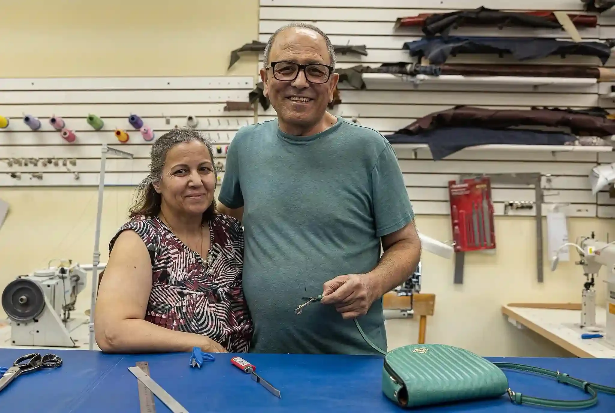 Smiling woman and man in a workshop or tailor shop, surrounded by fabric rolls on shelves, with sewing tools and a blue work table in front.