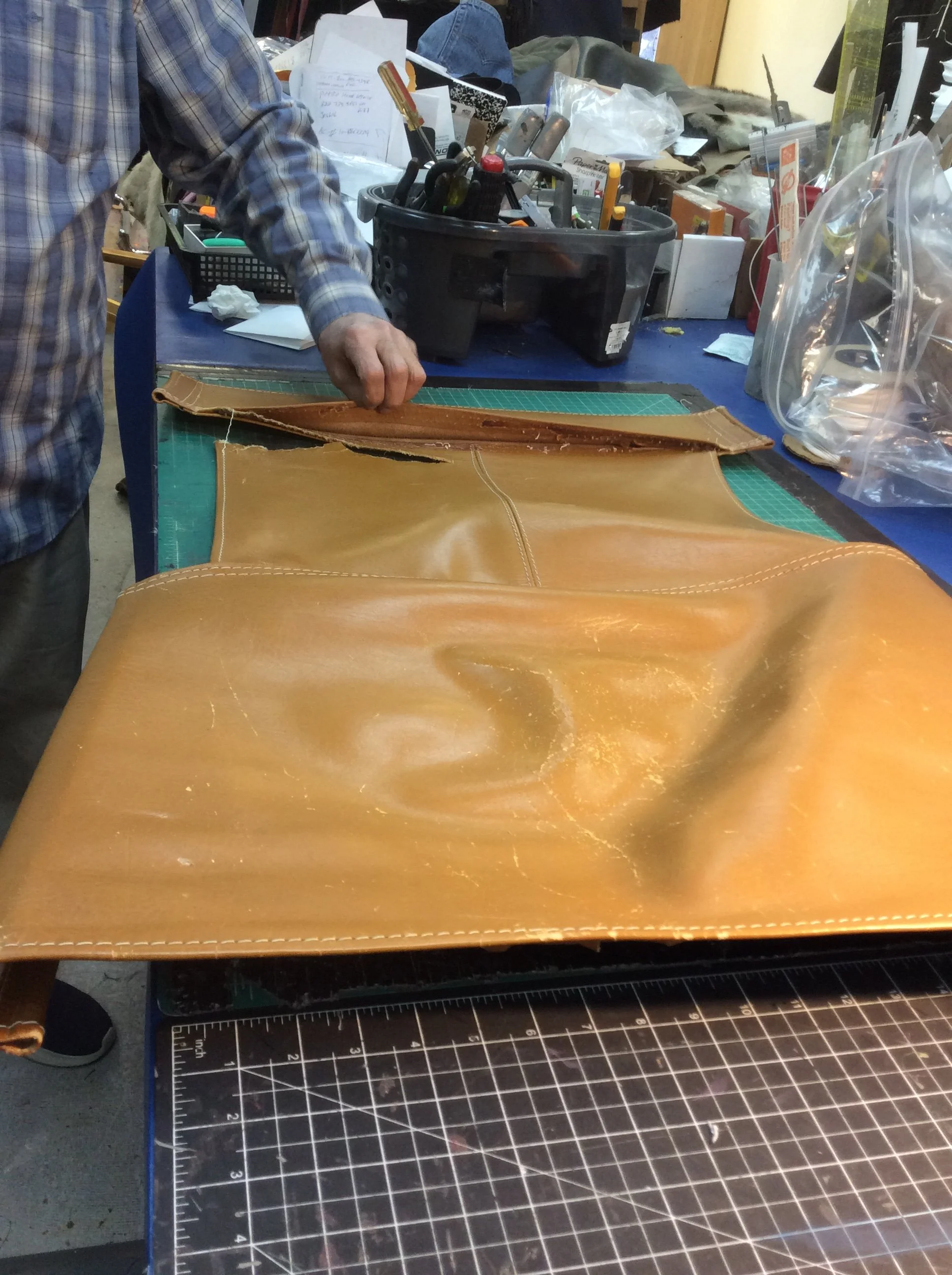 A person working on a leather project, cutting or shaping a piece of brown leather on a worktable, with various tools and supplies in the background.
