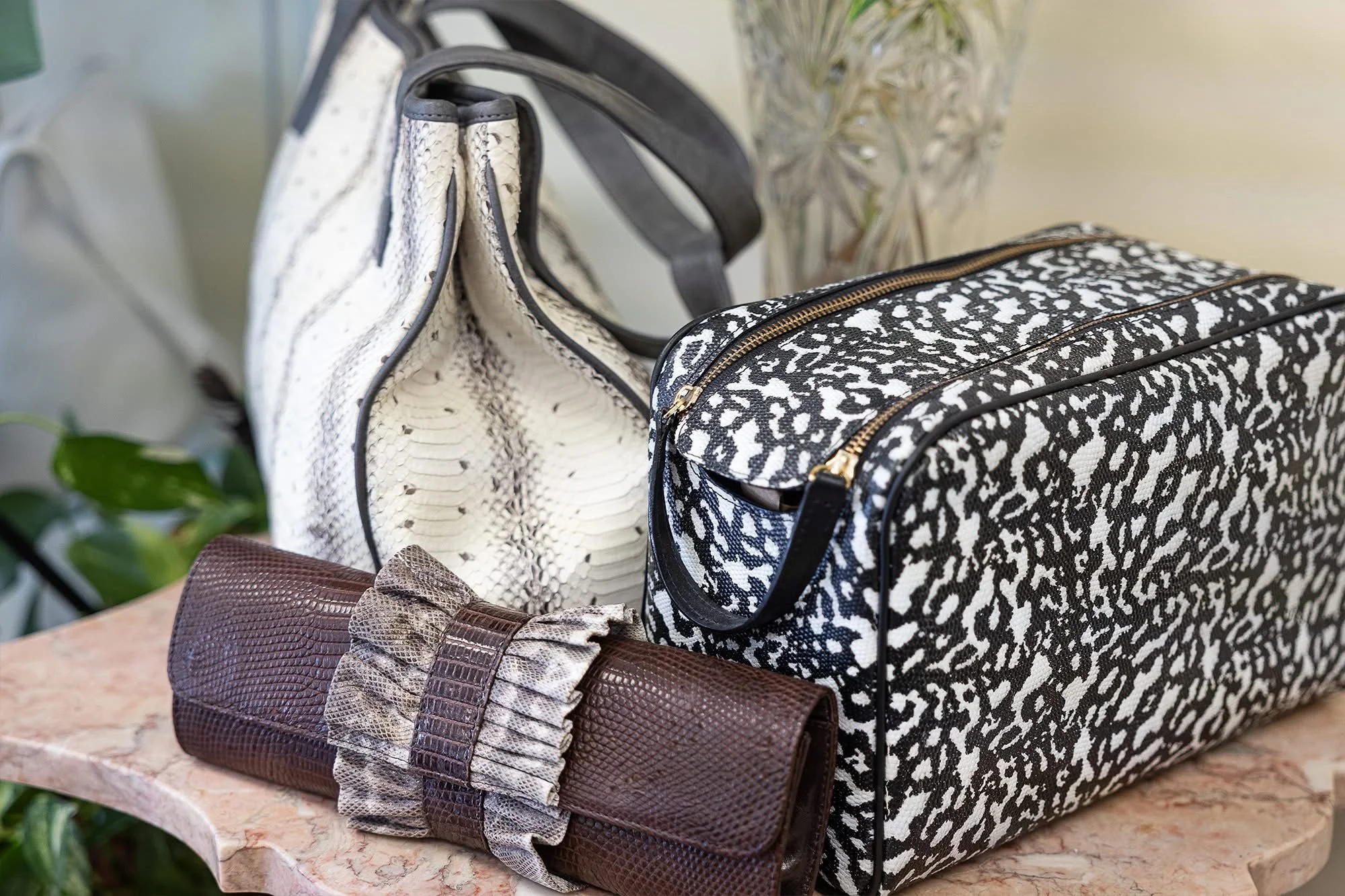 Collection of handbags made from snakeskin and animal print patterns displayed on a marble surface with a blurred background of green plants and a glass vase.