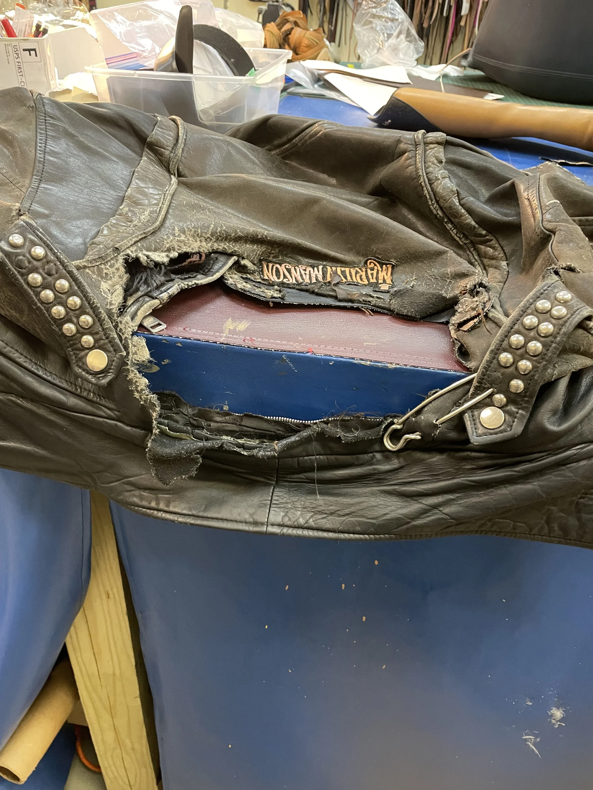 A worn, damaged black leather Harley Davidson jacket with studs on the cuffs, lying on a blue work surface. The background includes various items such as plastic containers, a wooden box, papers, and other objects, indicating a workshop or repair space.