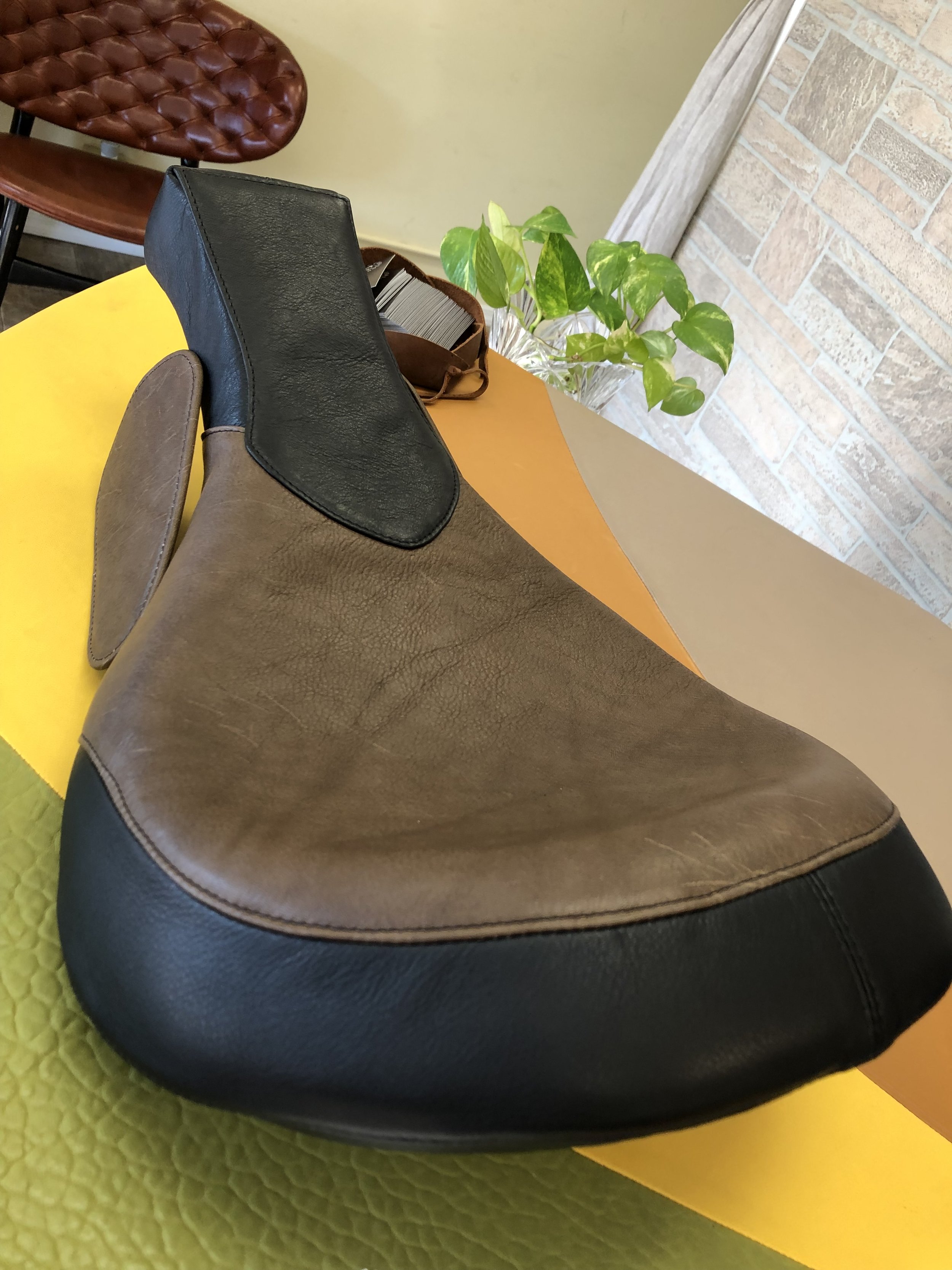 Close-up of a brown and black massage chair with a pair of brown and black cushions on it, set on a yellow textured surface, with a green potted plant in the background and a brick wall.