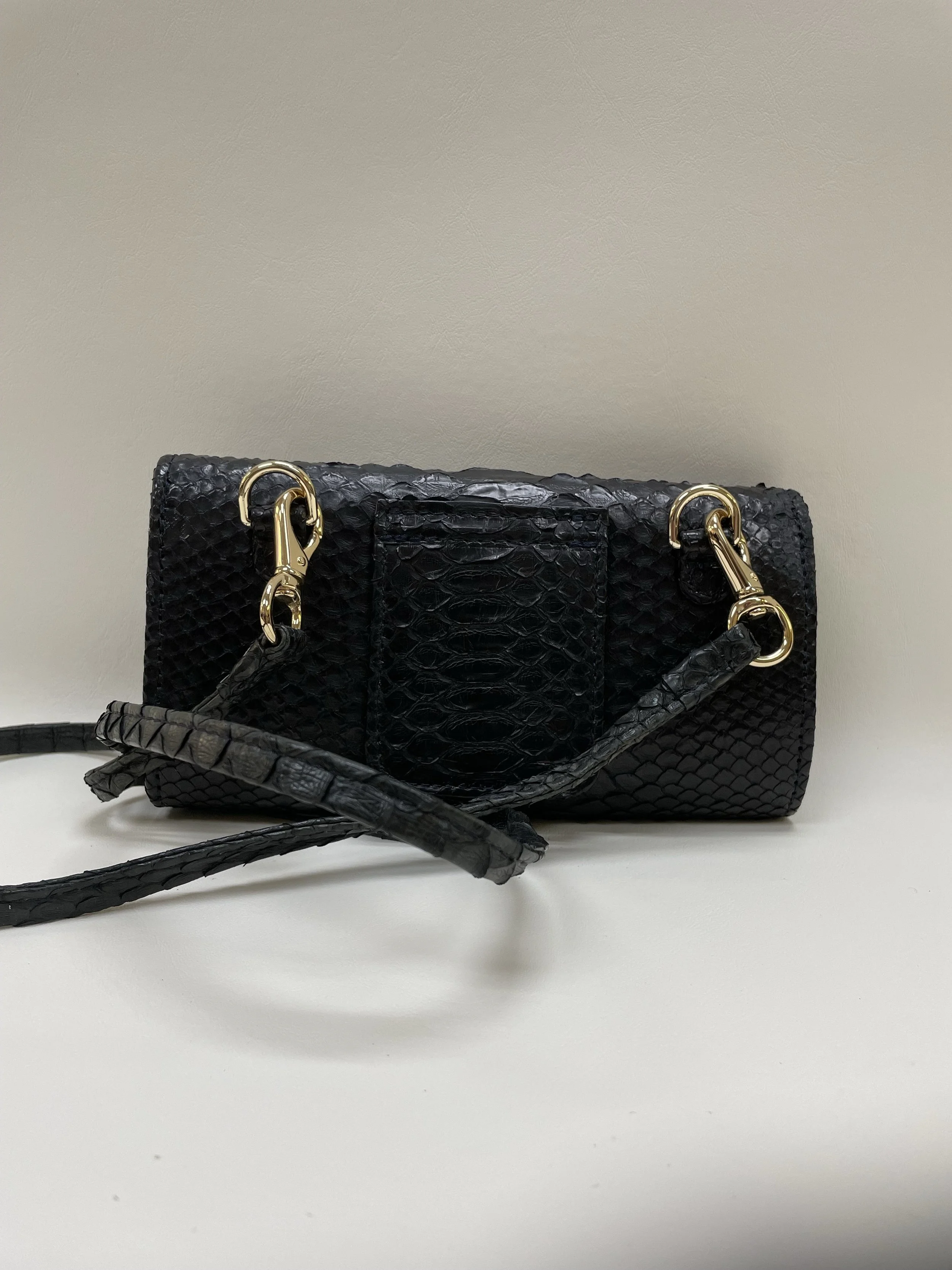 Close-up of a black handbag with a textured, snake-skin pattern and gold hardware, resting on a white surface with a plain light-colored background.