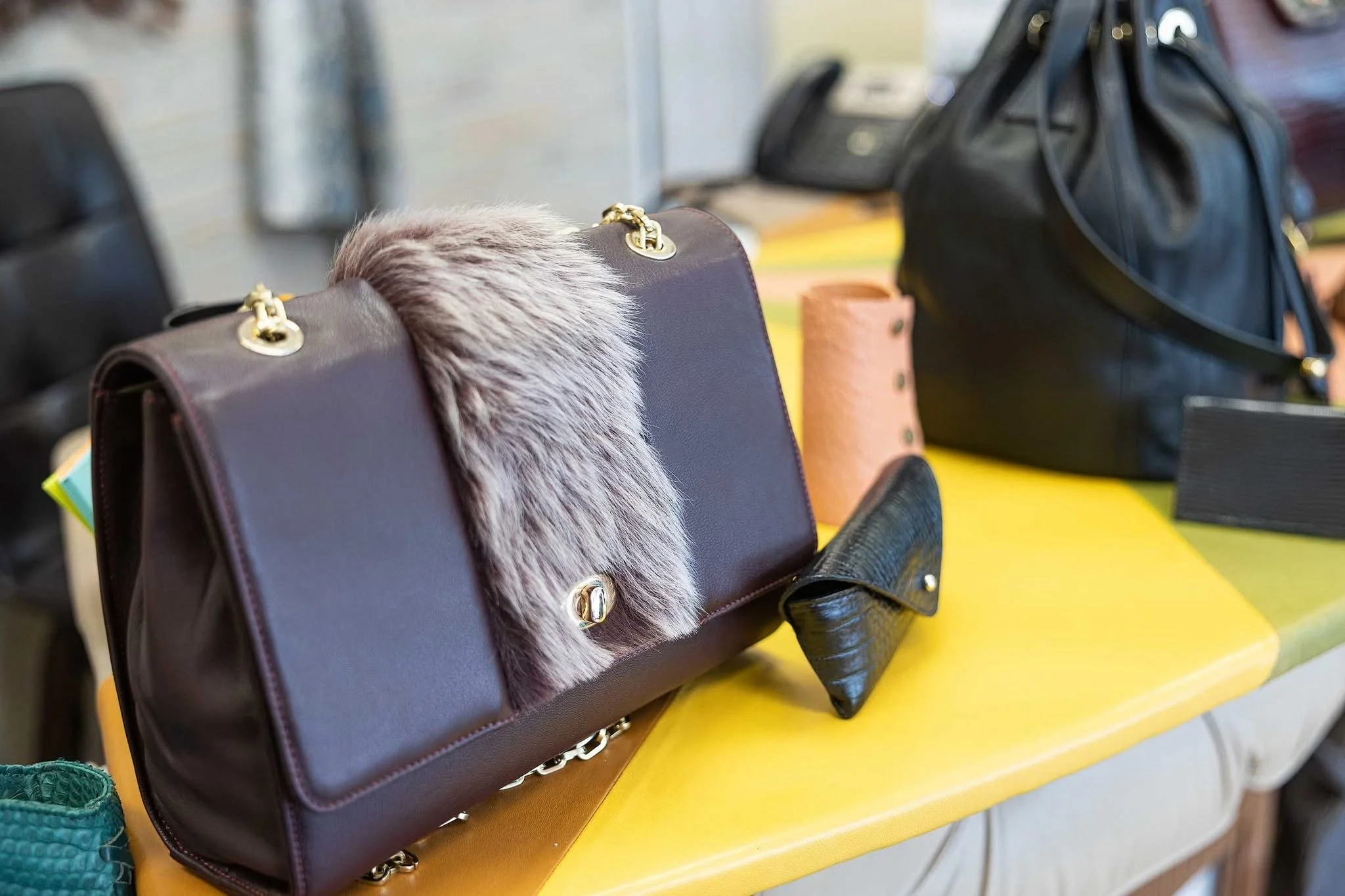 A collection of handbags and accessories displayed on a yellow table at a store or market. The handbags vary in colors and styles, with one featuring faux fur accents and gold hardware.
