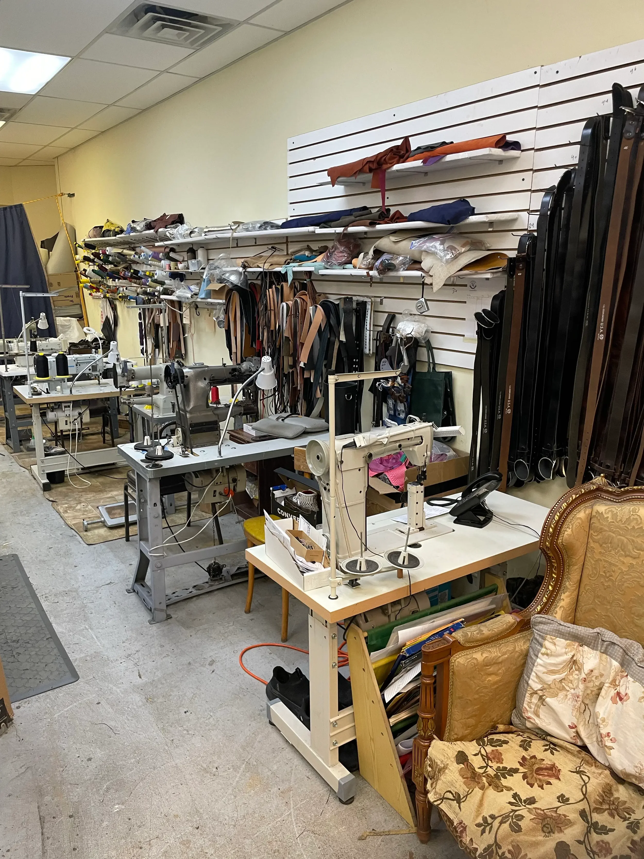 A cluttered workspace with sewing machines, fabric rolls, and various sewing supplies, along with a vintage floral upholstered armchair with pillows.