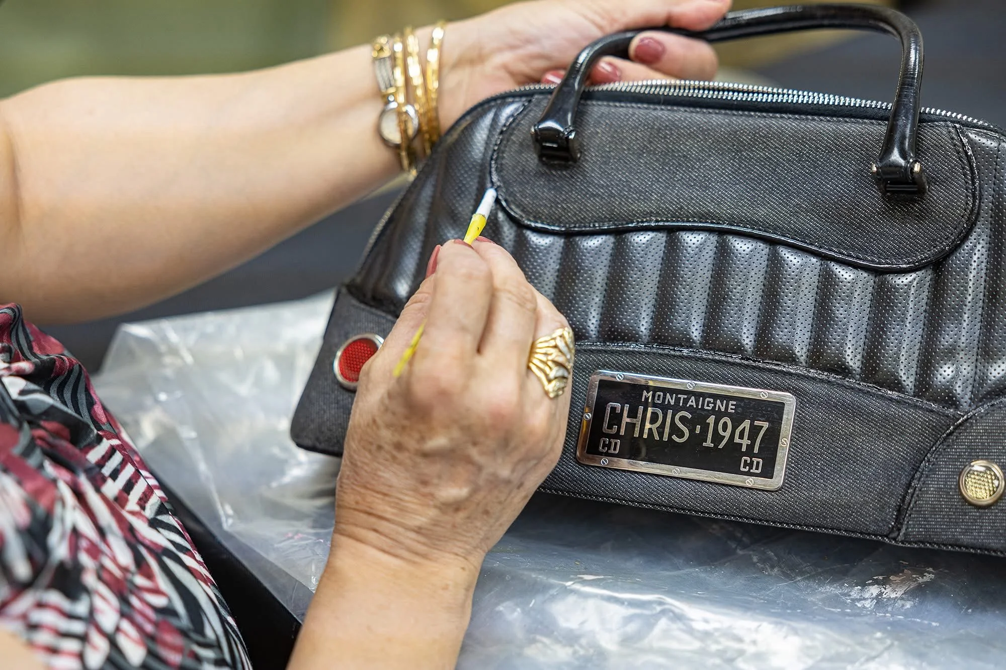 Close-up of a person's hands attaching a yellow cable to a black handbag with a license plate label reading 'Montaigne CHRIS 1947.'