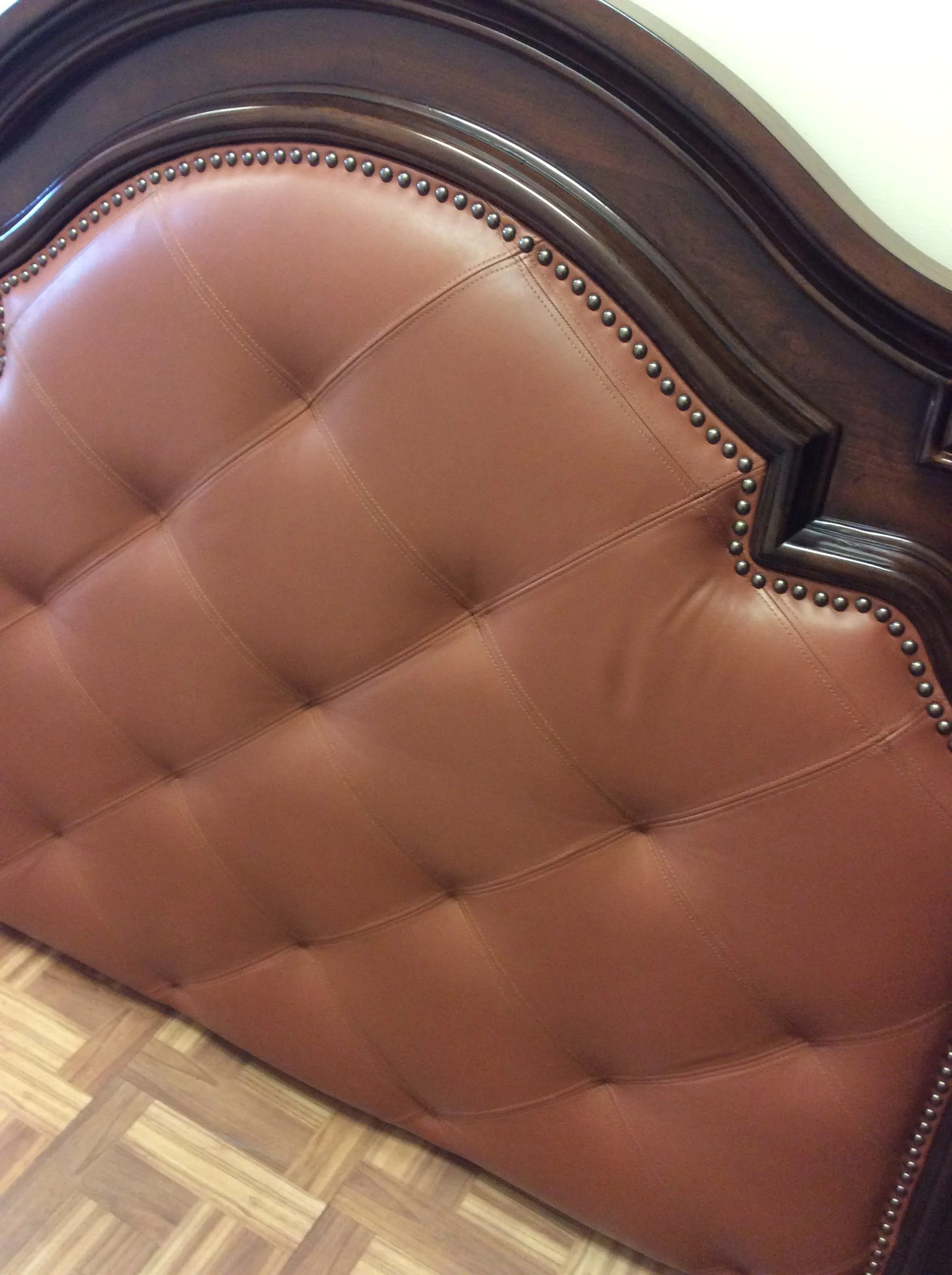 Close-up of a brown leather padded headboard with decorative nail head trim, adjacent to a wooden floor.