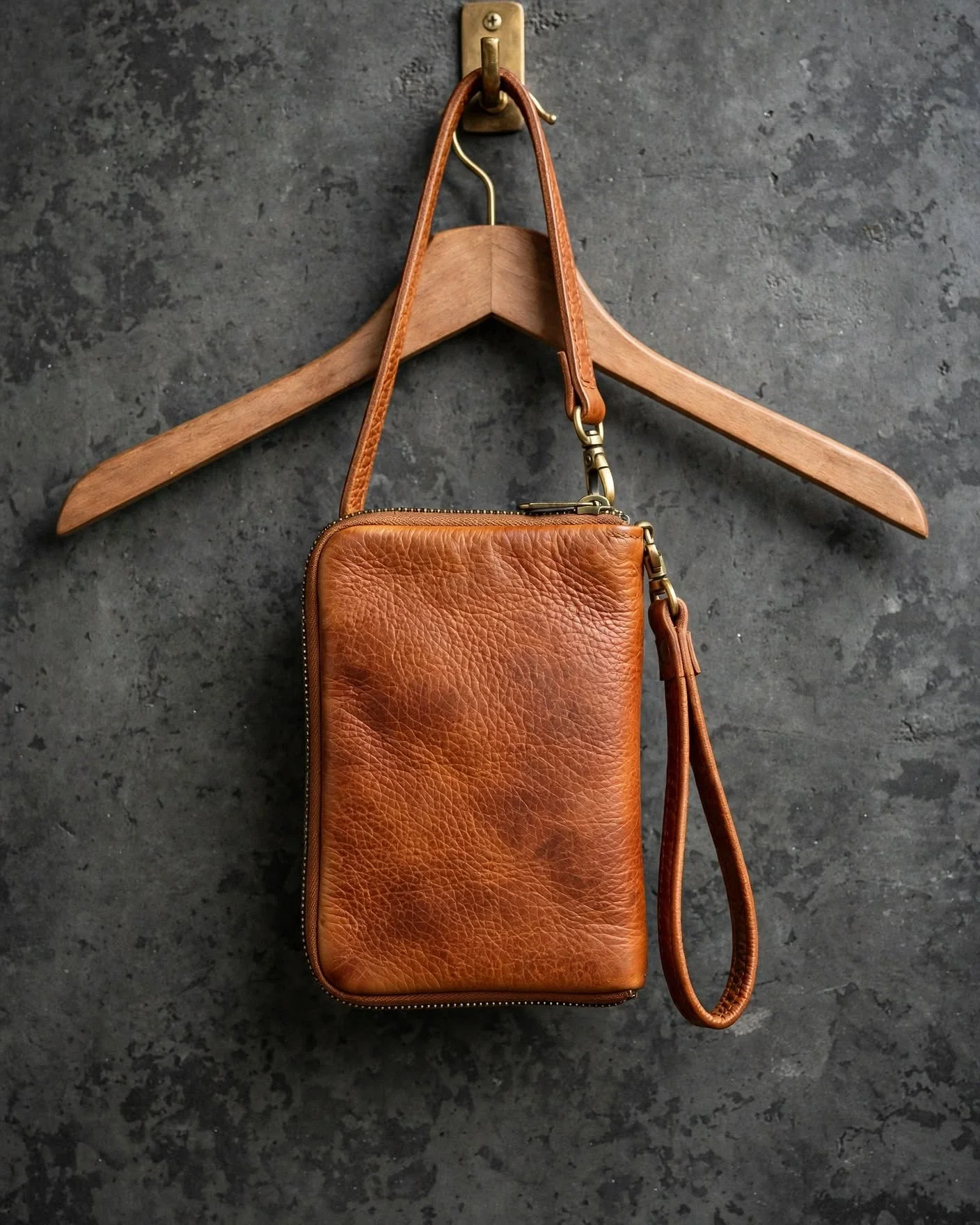 Natural tan pebbled leather wristlet with zip-around closure and detachable wrist strap hanging against dark charcoal wall