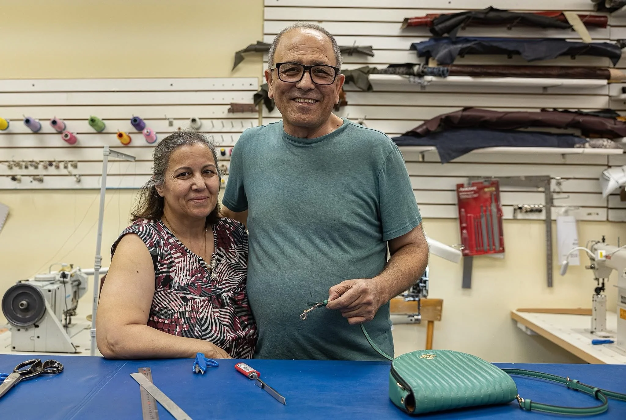 Two smiling people, one man and one woman, standing in a Leather Repair shop with fabric rolls on shelves behind them. The woman is wearing a patterned top, and the man is in a teal t-shirt and glasses.