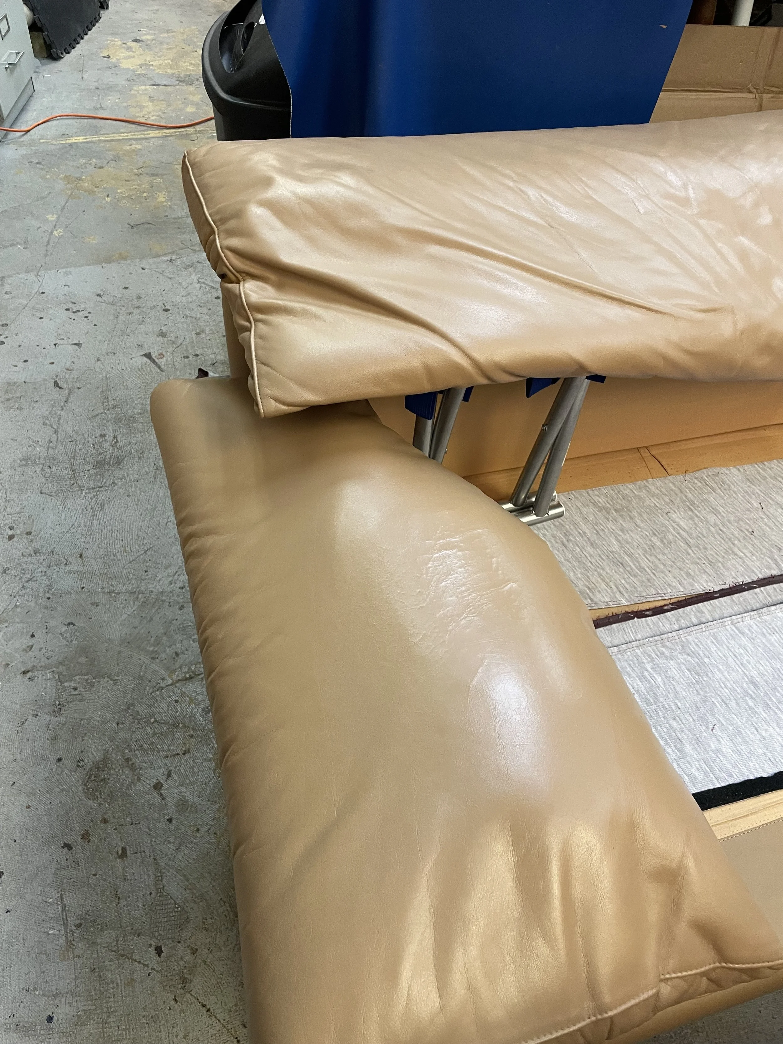A beige-colored padded folding massage table or bed with a diaper-like cover, situated on a concrete floor in a workshop or storage area.