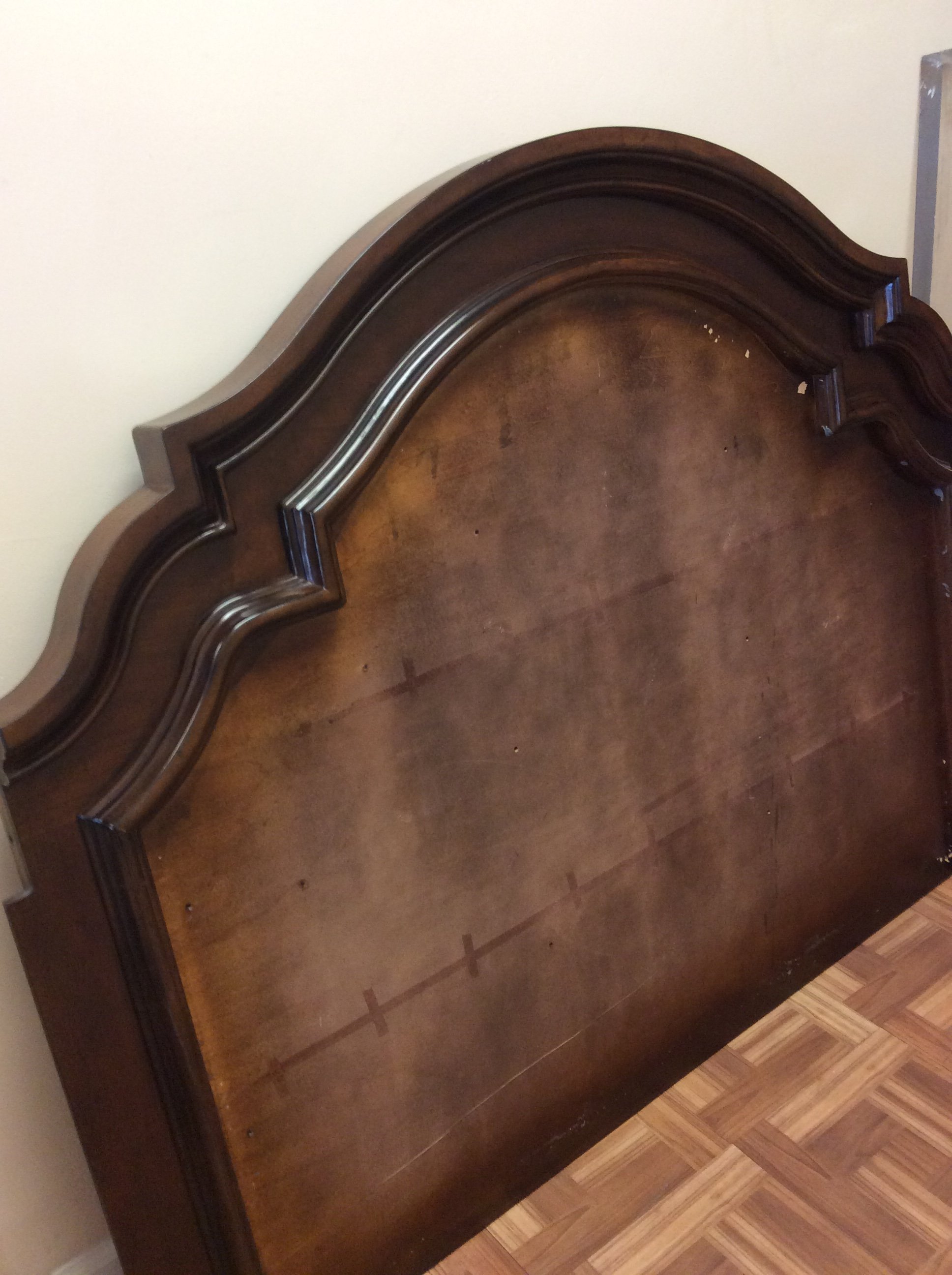Close-up of a dark wooden headboard with carved details and some visible wear, against a light-colored wall, with a portion of a patterned wooden floor visible at the bottom.