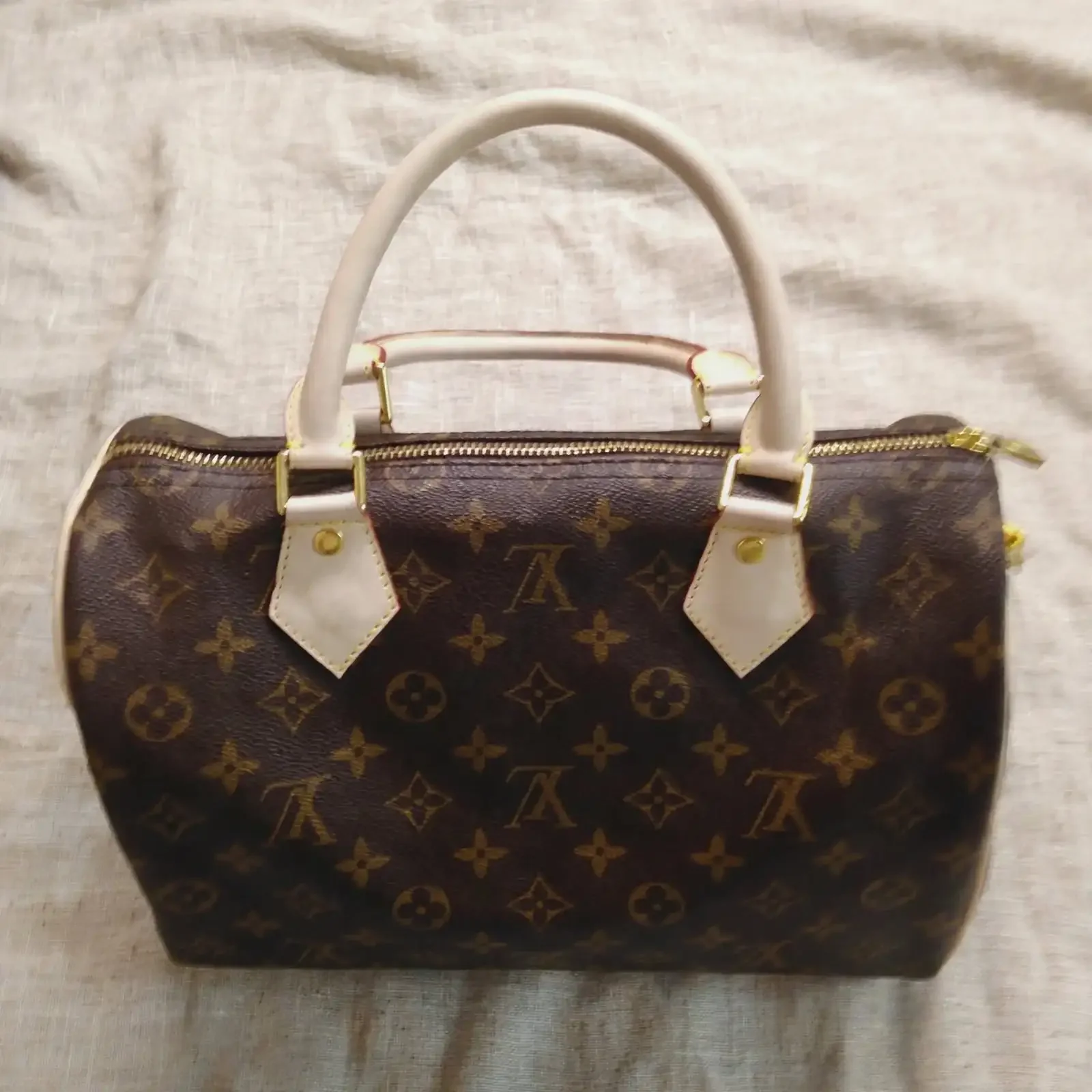 A designer handbag with Louis Vuitton monogram pattern, beige handles, and a gold zipper sitting on a light-colored fabric surface.