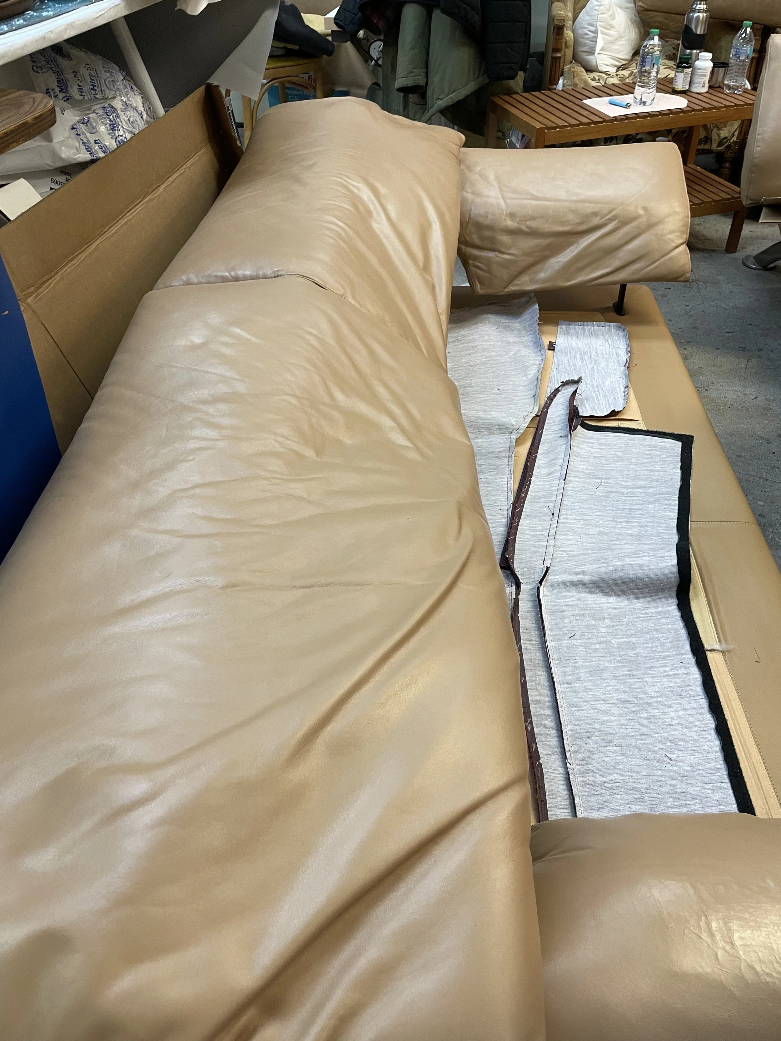 A beige leather couch in a cluttered room with various items including water bottles, bottles of medication, a wooden table, and clothing hanging behind it.