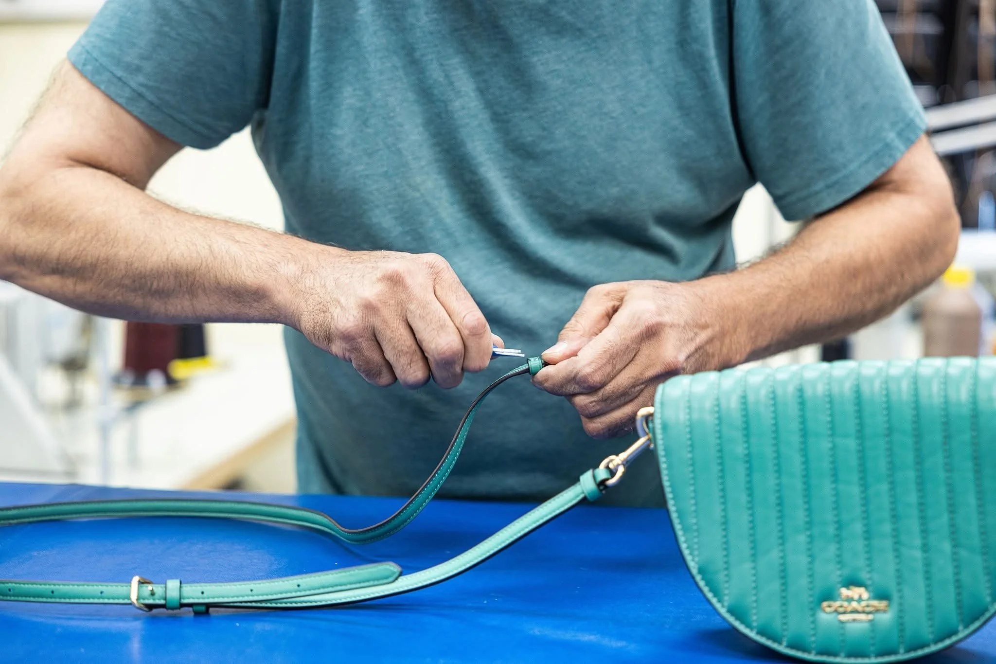 A person wearing a teal shirt working with electronic equipment, handling a wire connector, with a teal handbag and industrial-looking background.