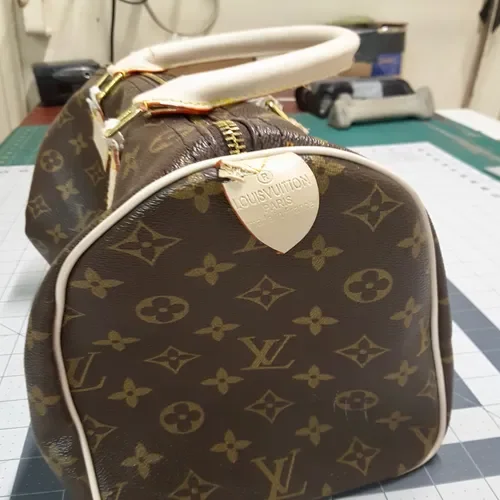 A Louis Vuitton handbag with monogram pattern, gold zipper, and white leather handles, placed on a table surface.