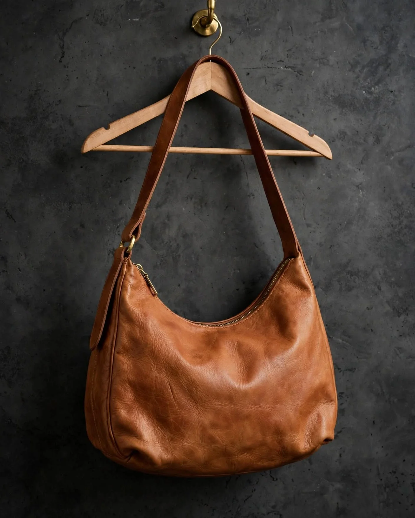 Natural tan leather hobo shoulder bag with crescent shape, zip closure and wide shoulder strap hanging on wooden hanger against dark concrete wall