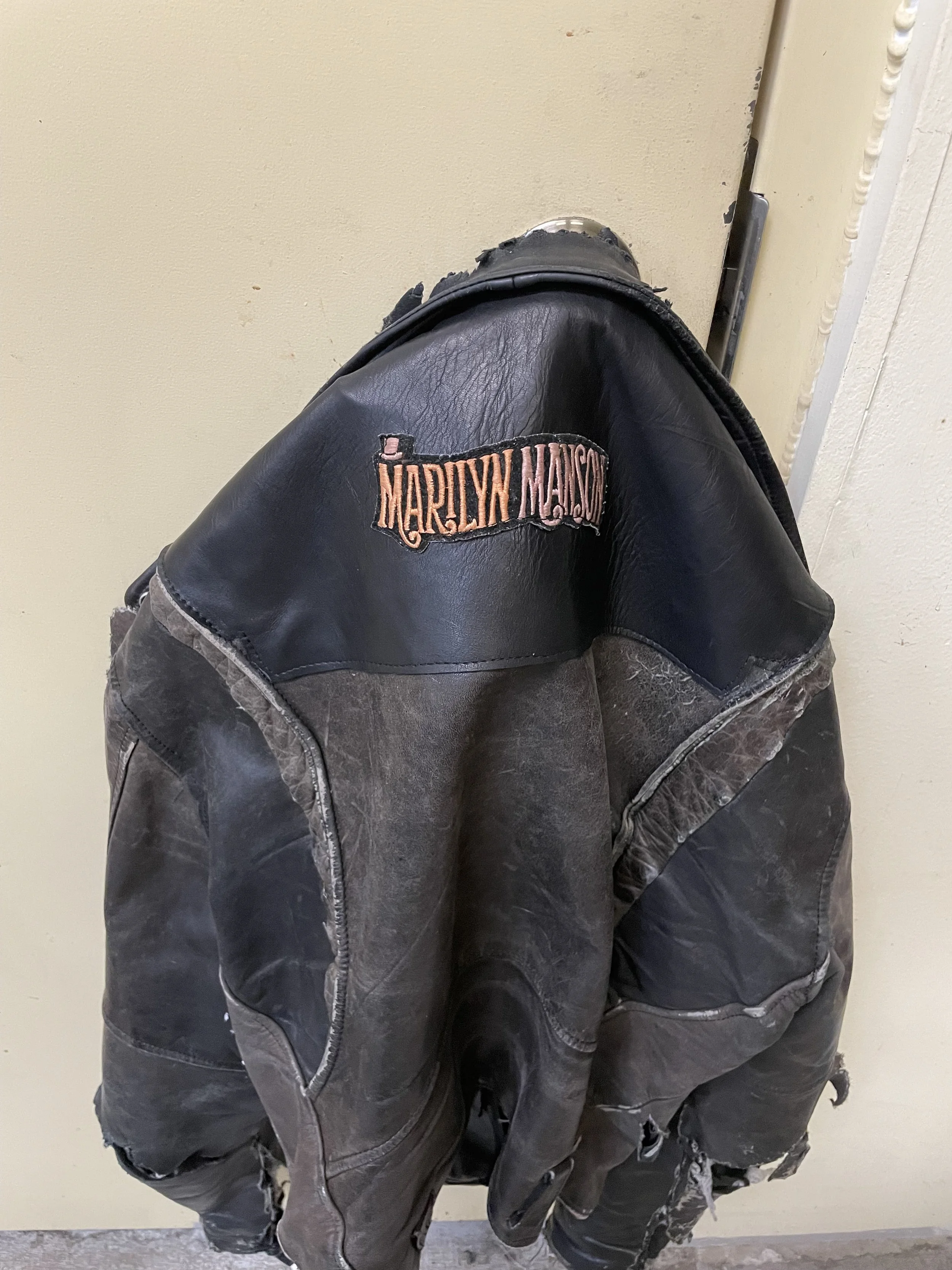 Close-up of a black leather Harley Davidson jacket hanging on a hook or hanger against a beige wall.