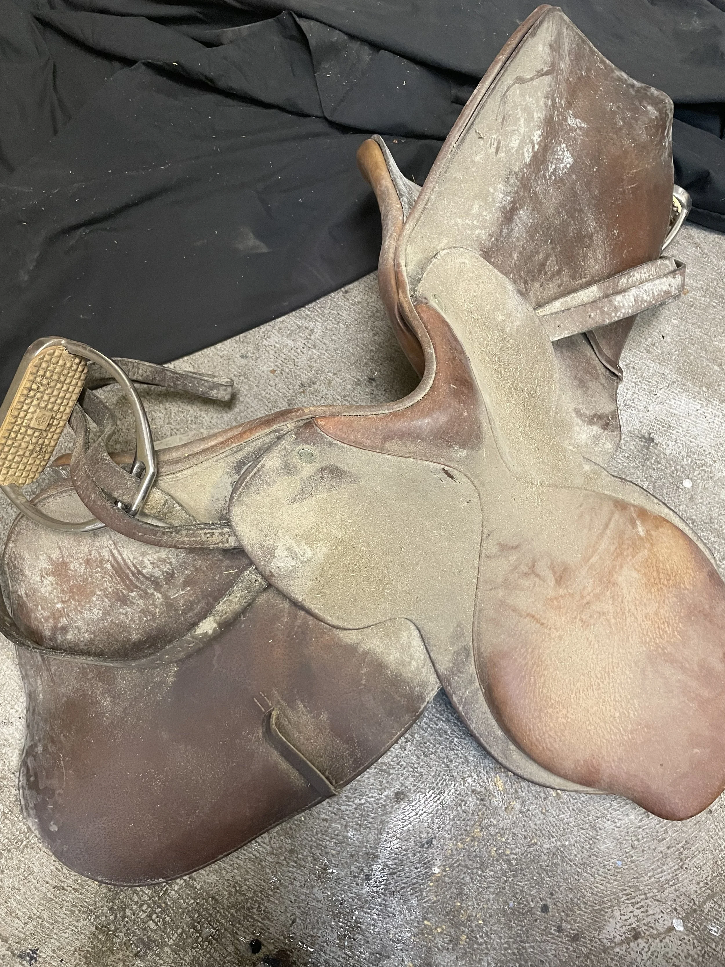 A collection of vintage saddles and a saddle tree placed on a textured surface, with some dust and dirt on the items.