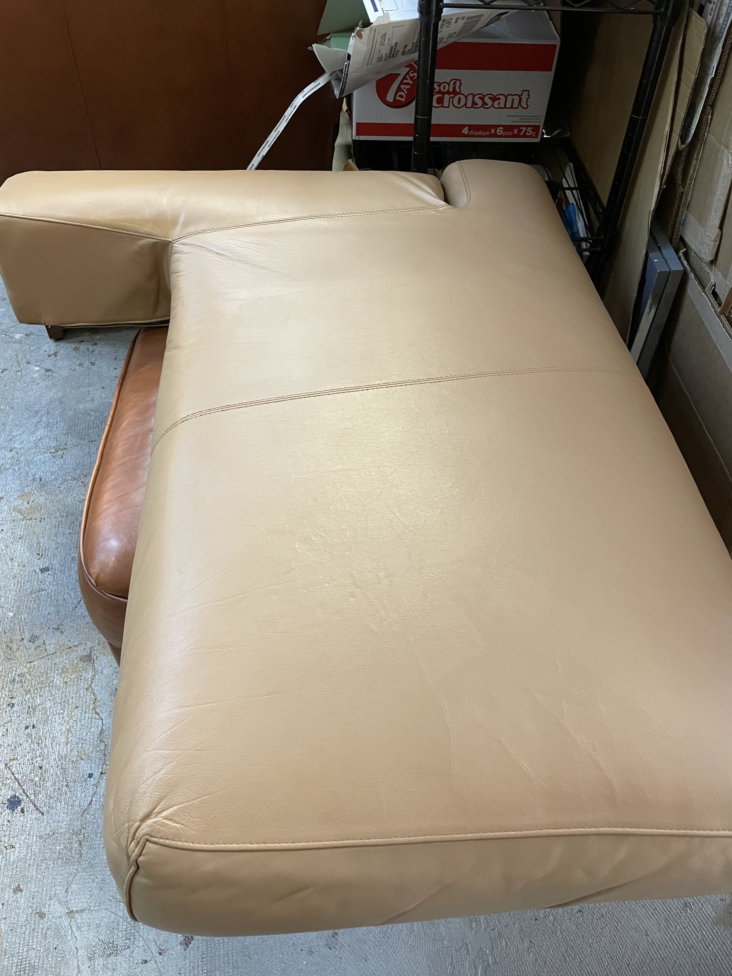 A beige or tan leather couch or lounge chair with a rounded armrest, situated on a concrete or unfinished floor in a storage or warehouse space.