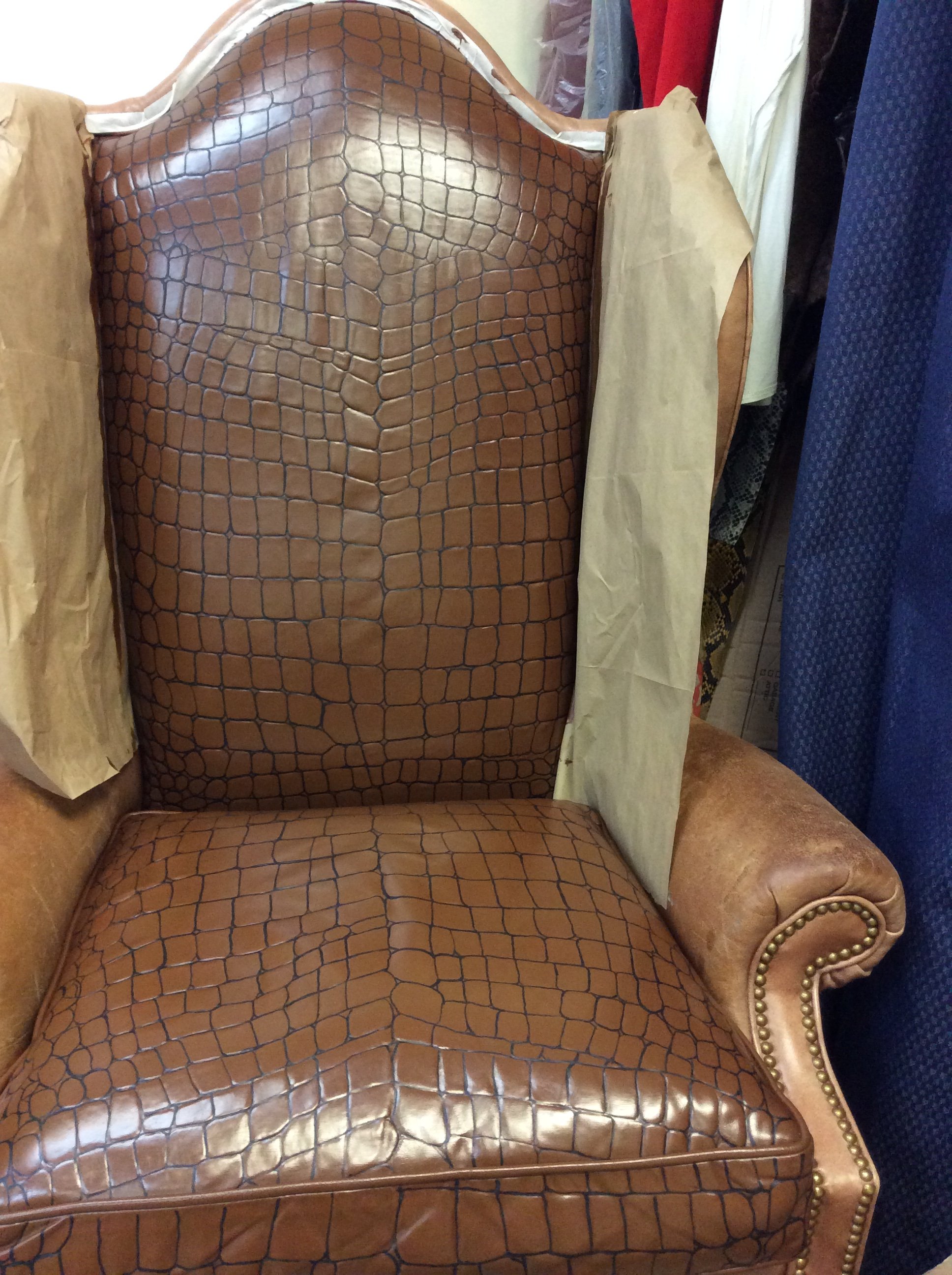 A vintage upholstered armchair with a crocodile skin pattern in tan and brown tones, featuring rounded arms with brass nailhead trim. There are fabric-covered cardboard cushions behind the backrest and a textured blue curtain in the background.