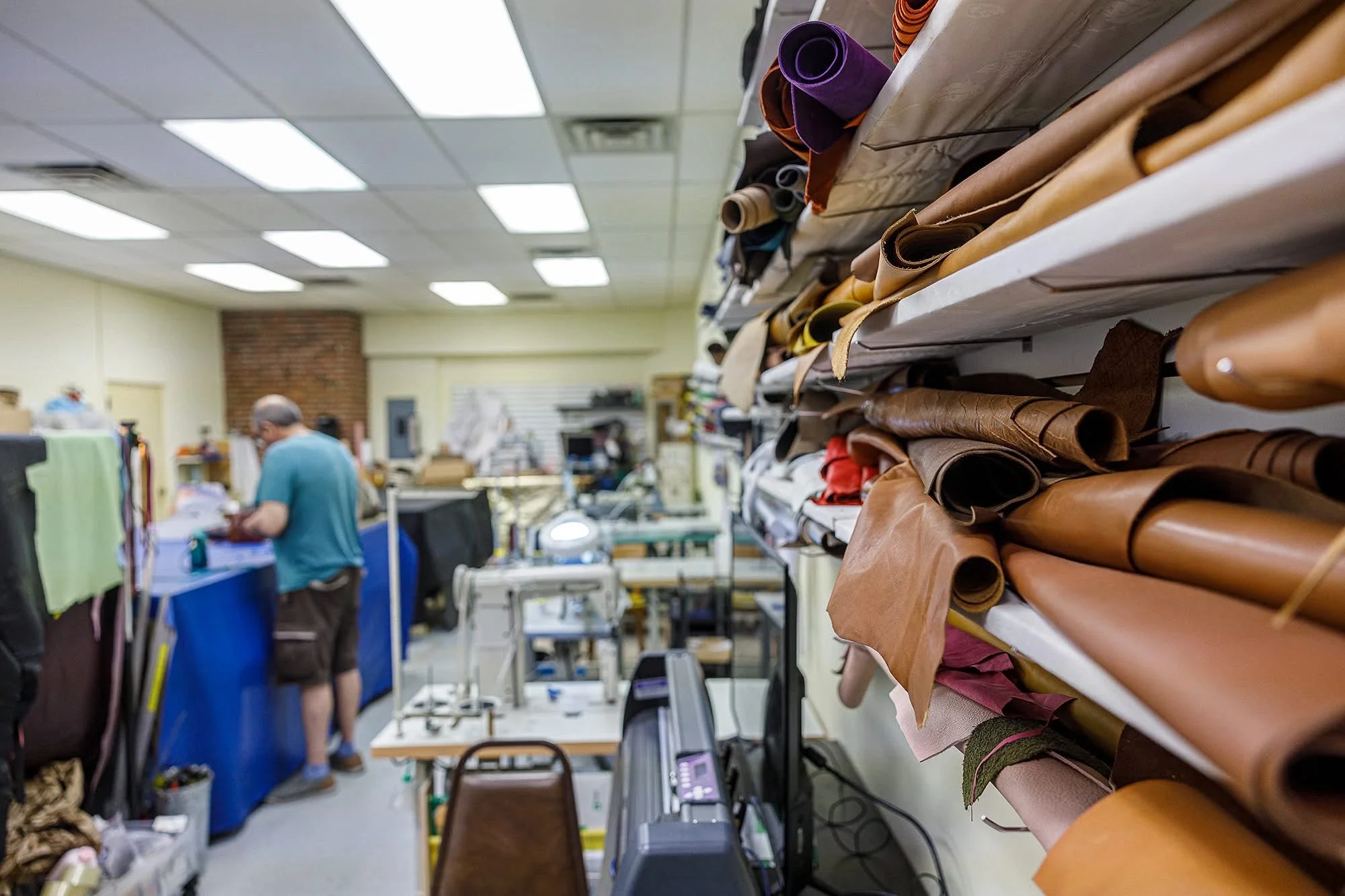 A person working in a leather repair workshop, standing at a table with leather rolls and leather supplies, with shelves of leather rolls in the foreground and ceiling lights overhead.