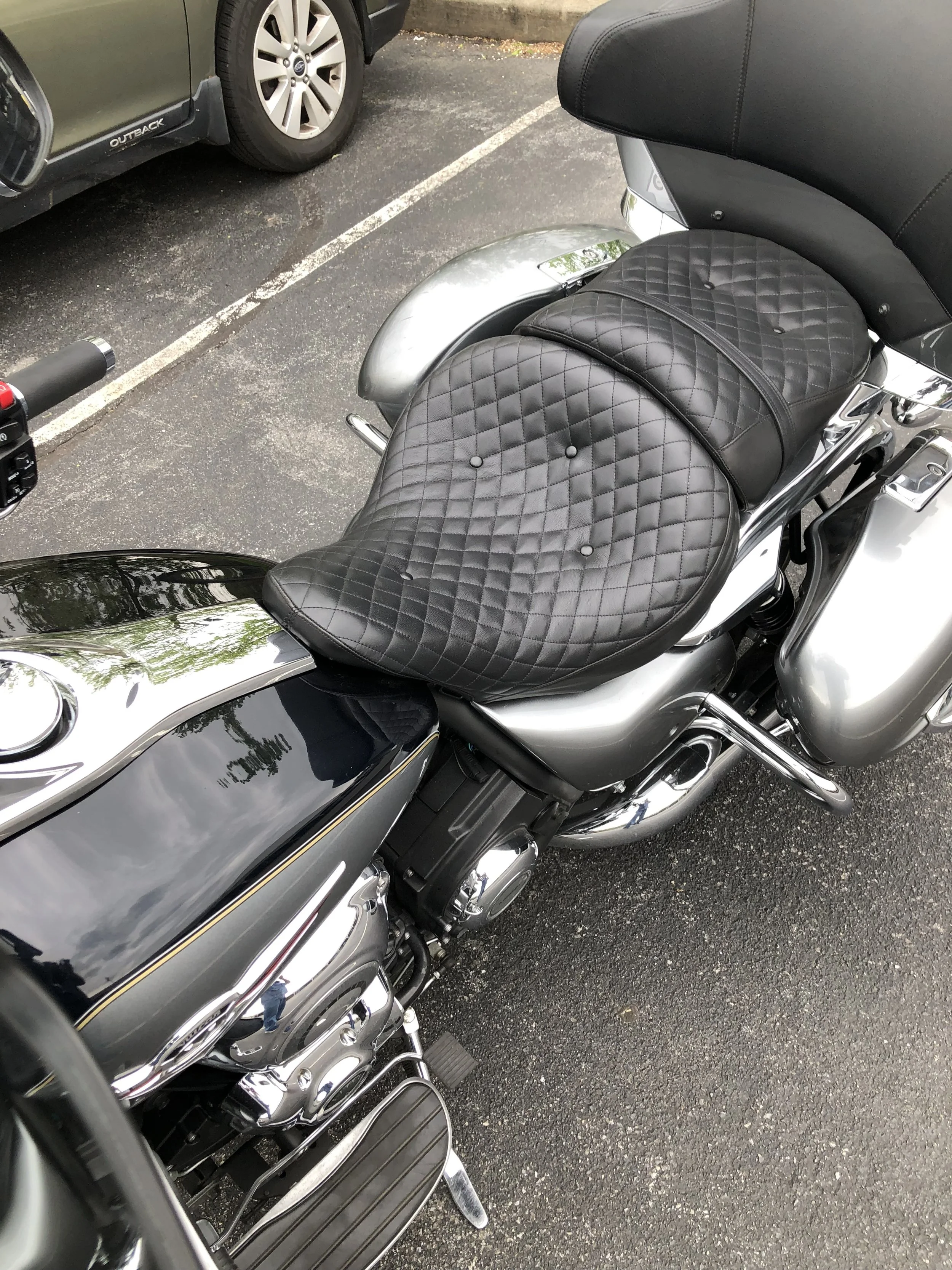 Close-up of a black motorcycle or scooter with quilted leather seat and chrome accents, parked on an asphalt lot next to a gray SUV.