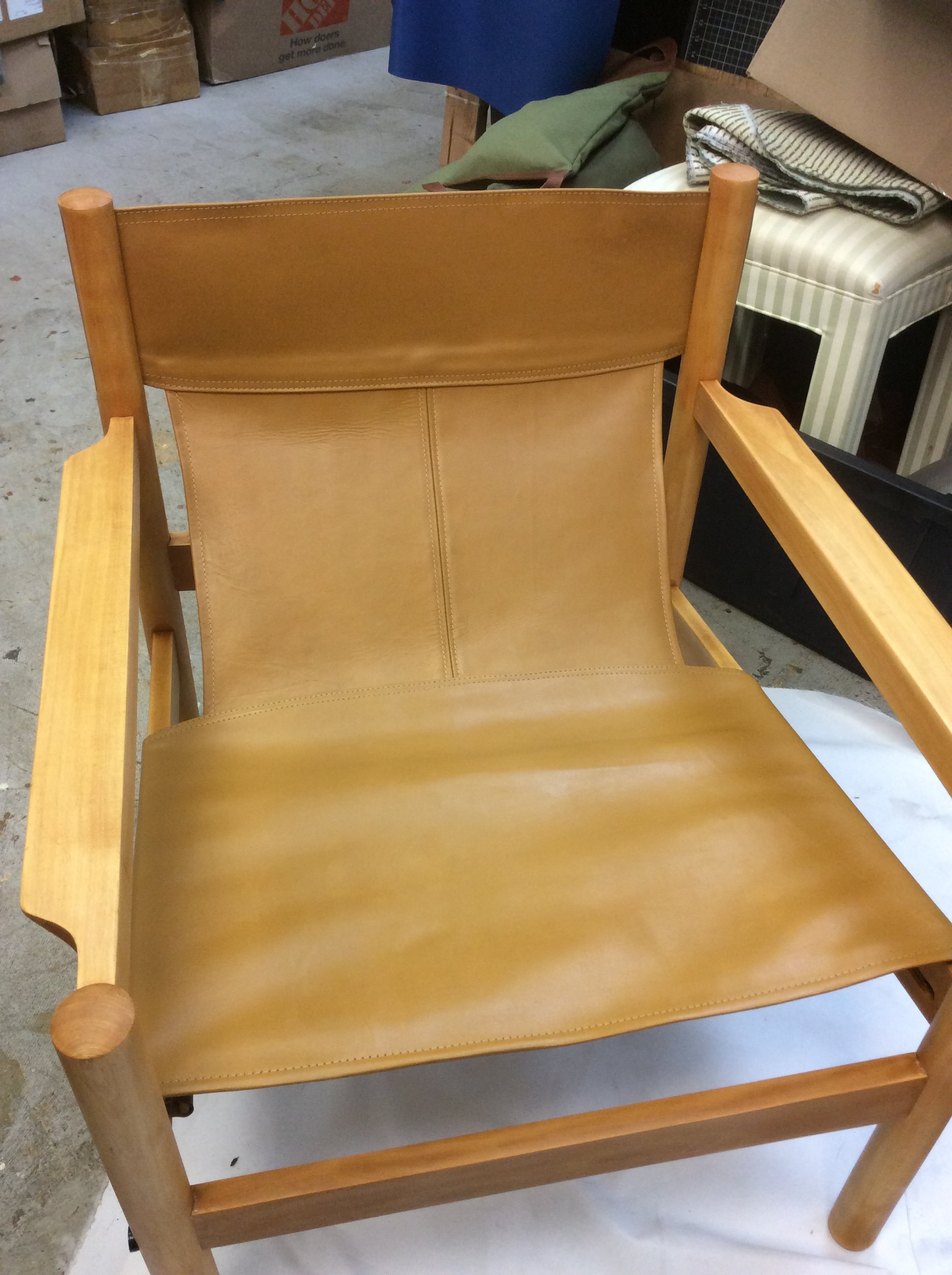 A wooden armchair with a cushioned tan leather backrest and seat, positioned in a garage or storage area with various boxes and furniture around.