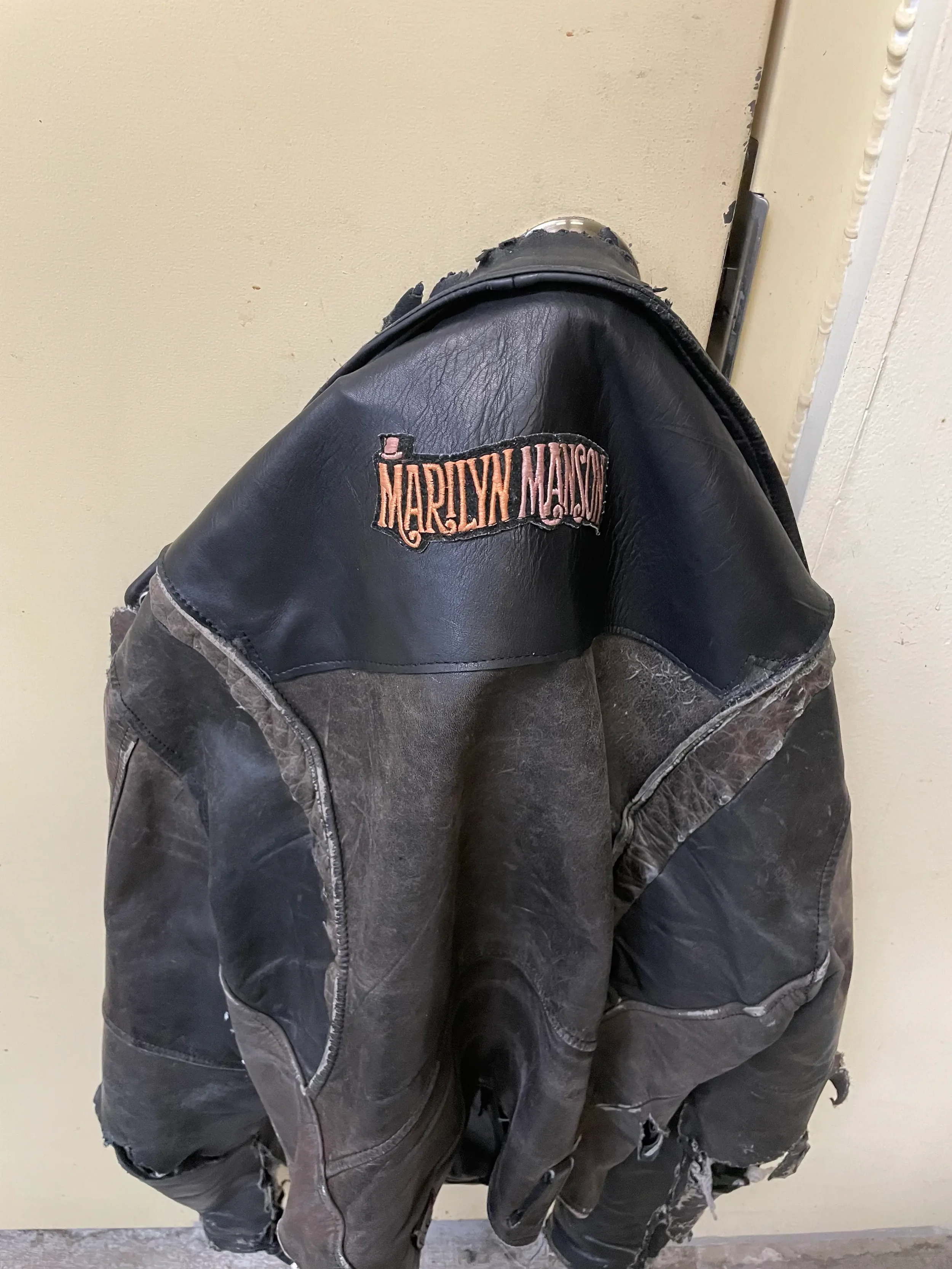 A worn black leather Harley Davidson jacket with a patch reading 'Marilyn Monroe' on the back, draped over a person's lap as they sit on a chair or bench against a plain beige wall.