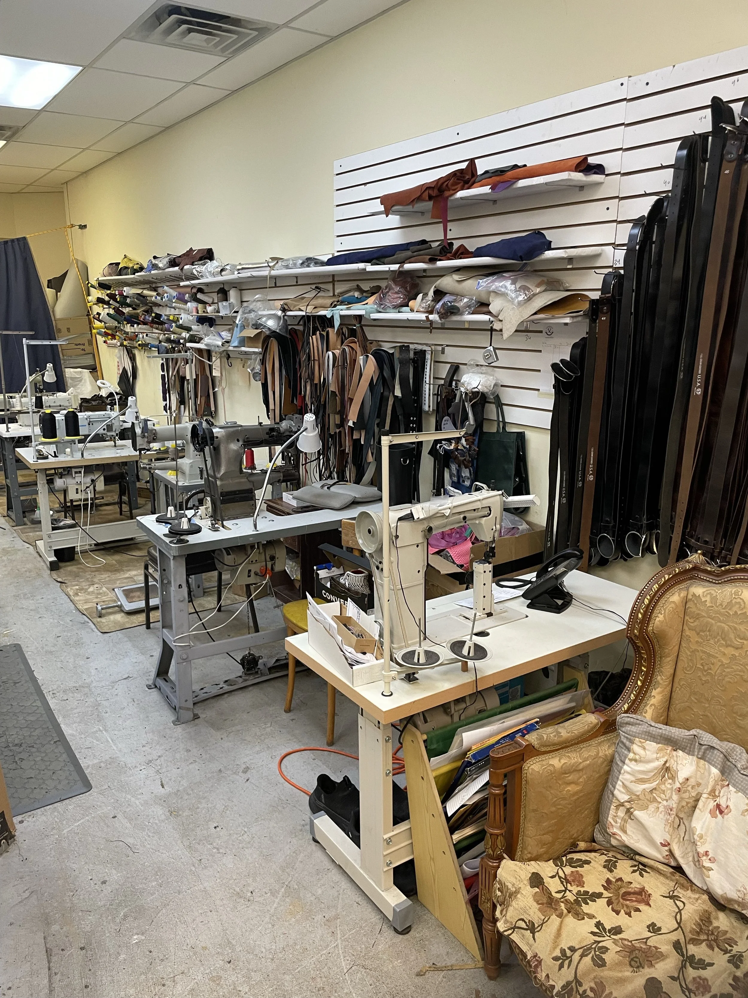 A cluttered workspace with sewing machines, fabric rolls, and various sewing tools. There's a vintage armchair with floral upholstery and pillows, a wall with white slats holding fabric and supplies, and fluorescent ceiling lights.